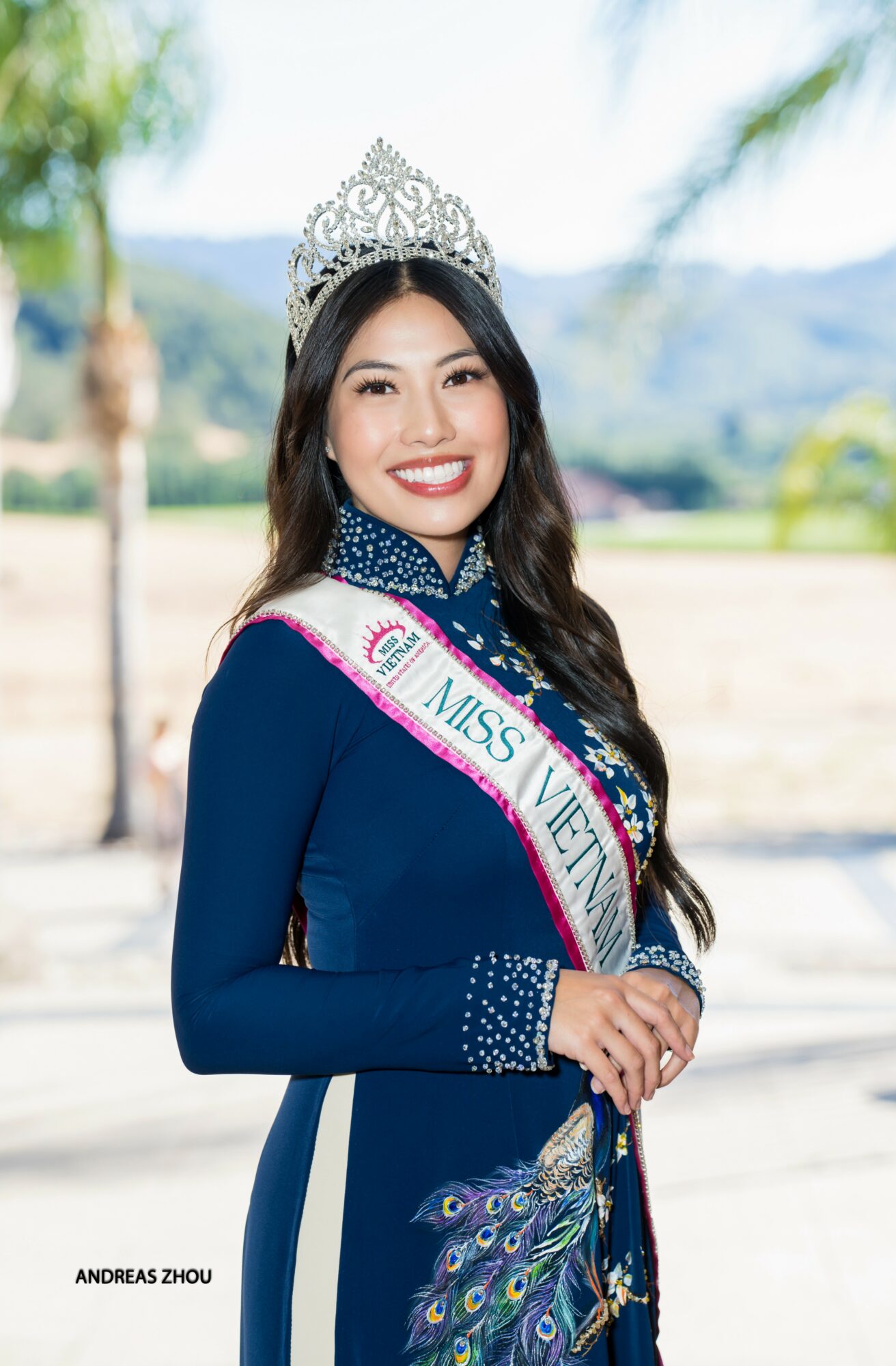 Woman wearing a crown and sash outdoors, smiling, with trees and mountains in background.