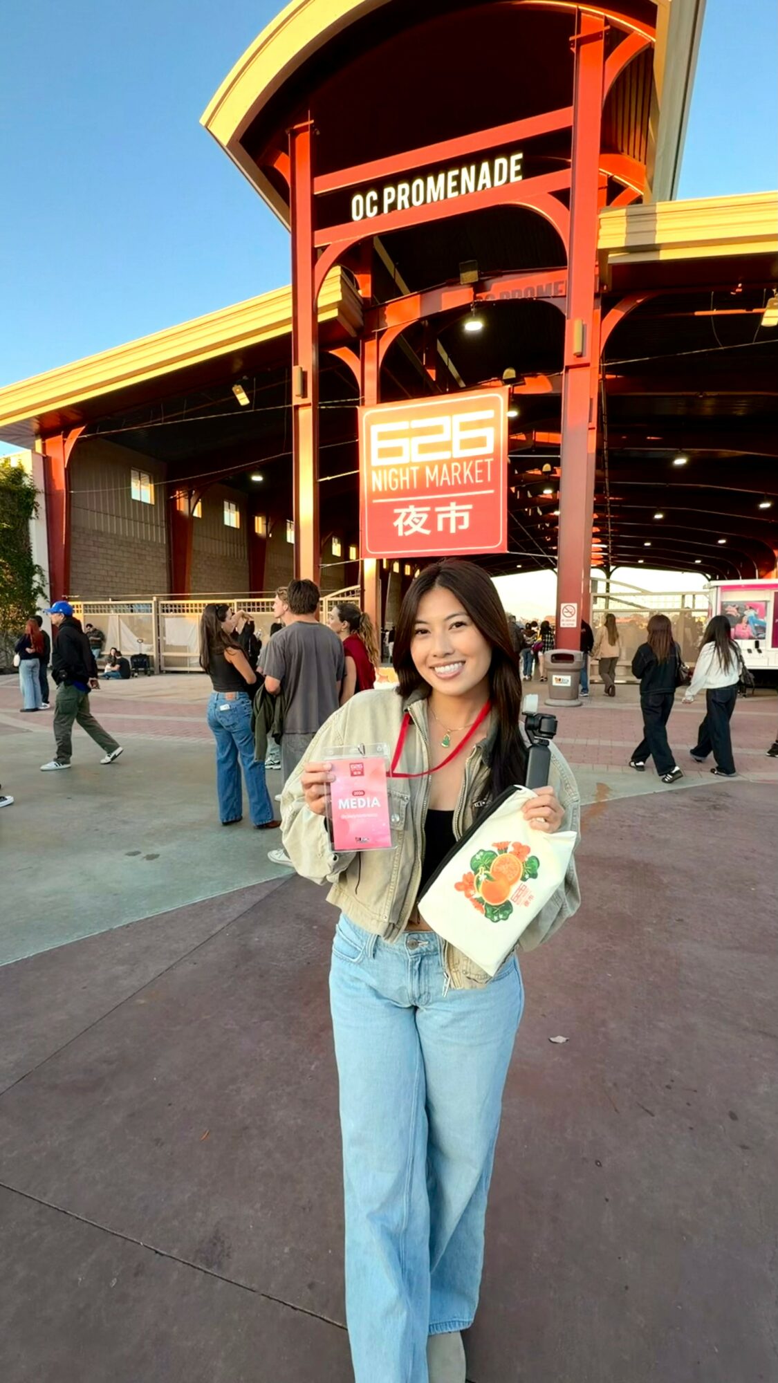 Young woman holding a drink and a bag, standing in front of a red and yellow entrance sign at OC Promenade.