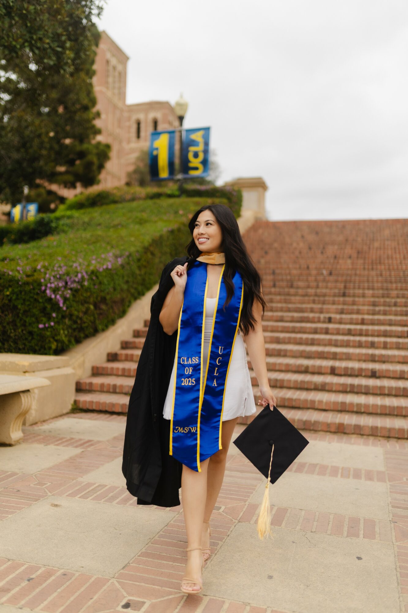 Young woman in graduation attire holding a diploma and a cap, standing on stairs outdoors with a building and greenery behind her.