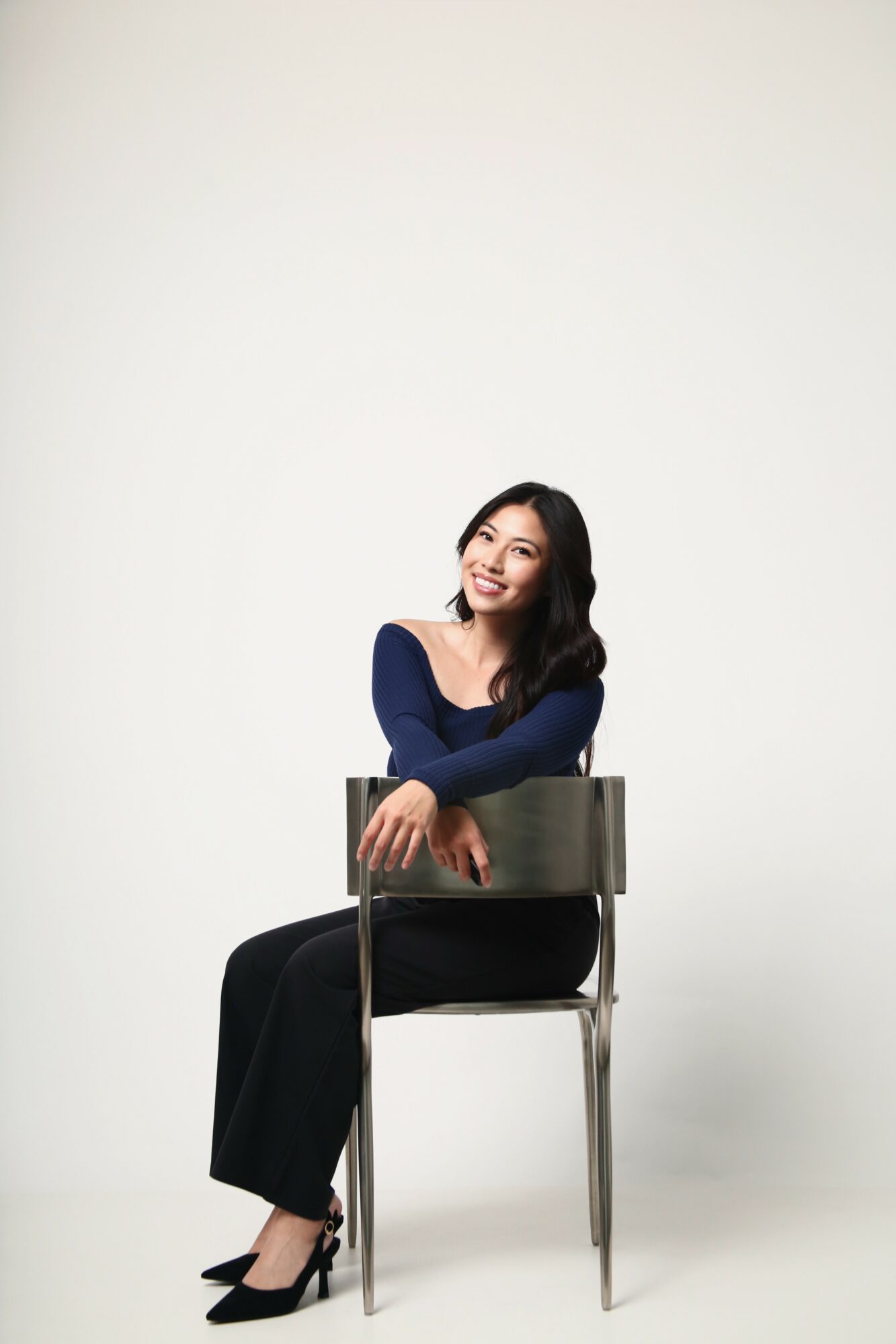 Woman sitting on a chair, smiling, with a plain white background.