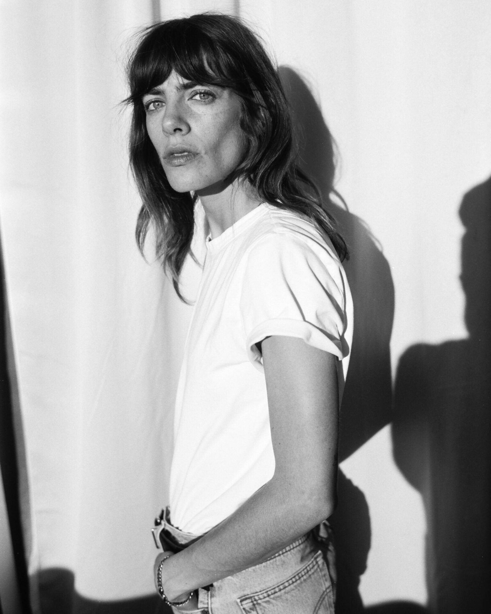 Woman with dark hair and bangs, wearing a white t-shirt, standing against a plain background, looking at the camera.