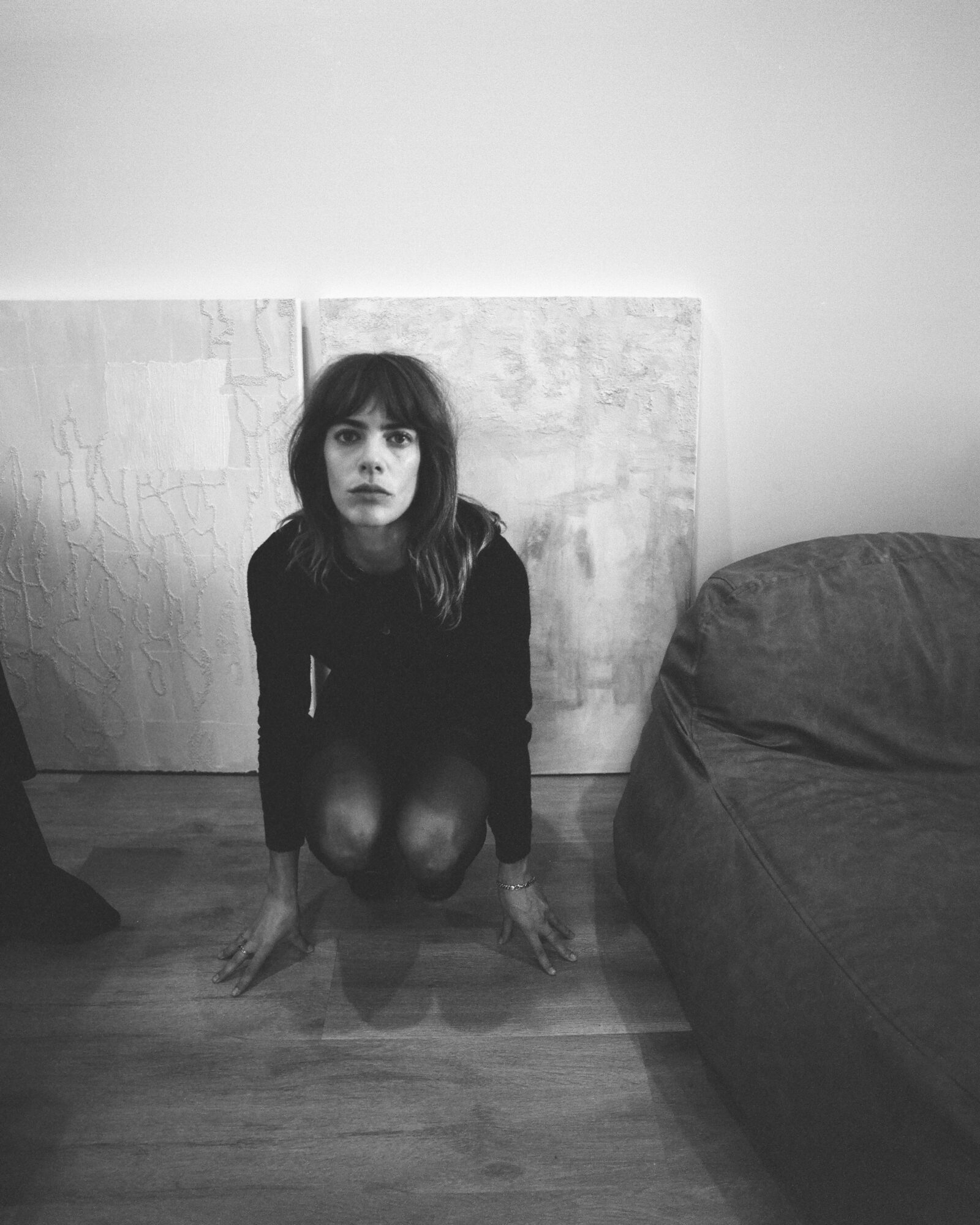 Woman with dark hair and bangs crouching on floor, looking up, next to a sofa and wall, in black and white.