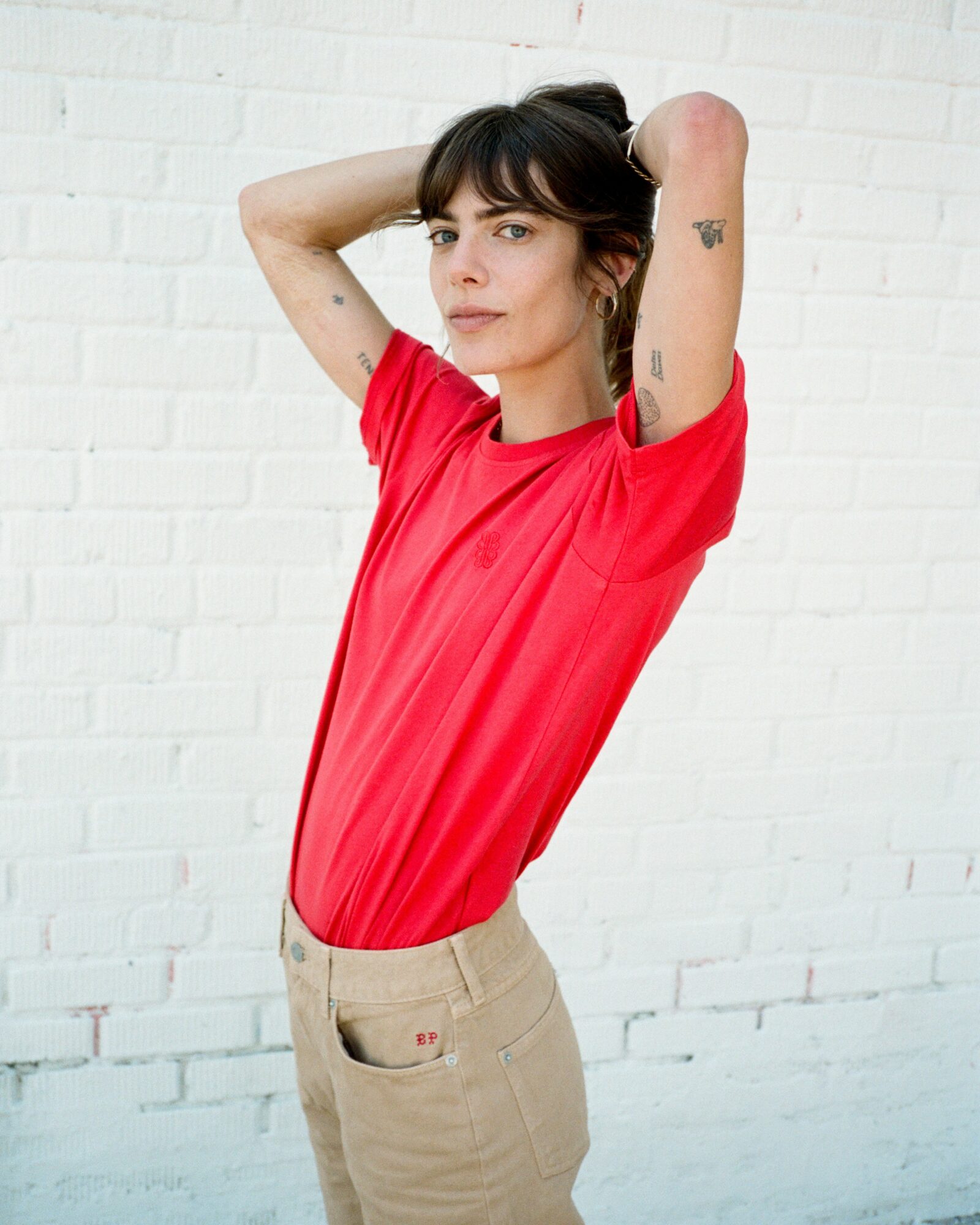 Young woman with dark hair and tattoos, wearing a red shirt and beige pants, standing against a white brick wall.