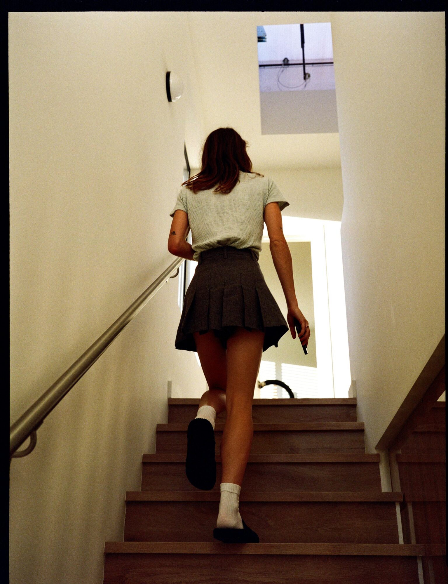 Person with long hair ascending stairs in a well-lit indoor space, viewed from behind, with a window at the top.