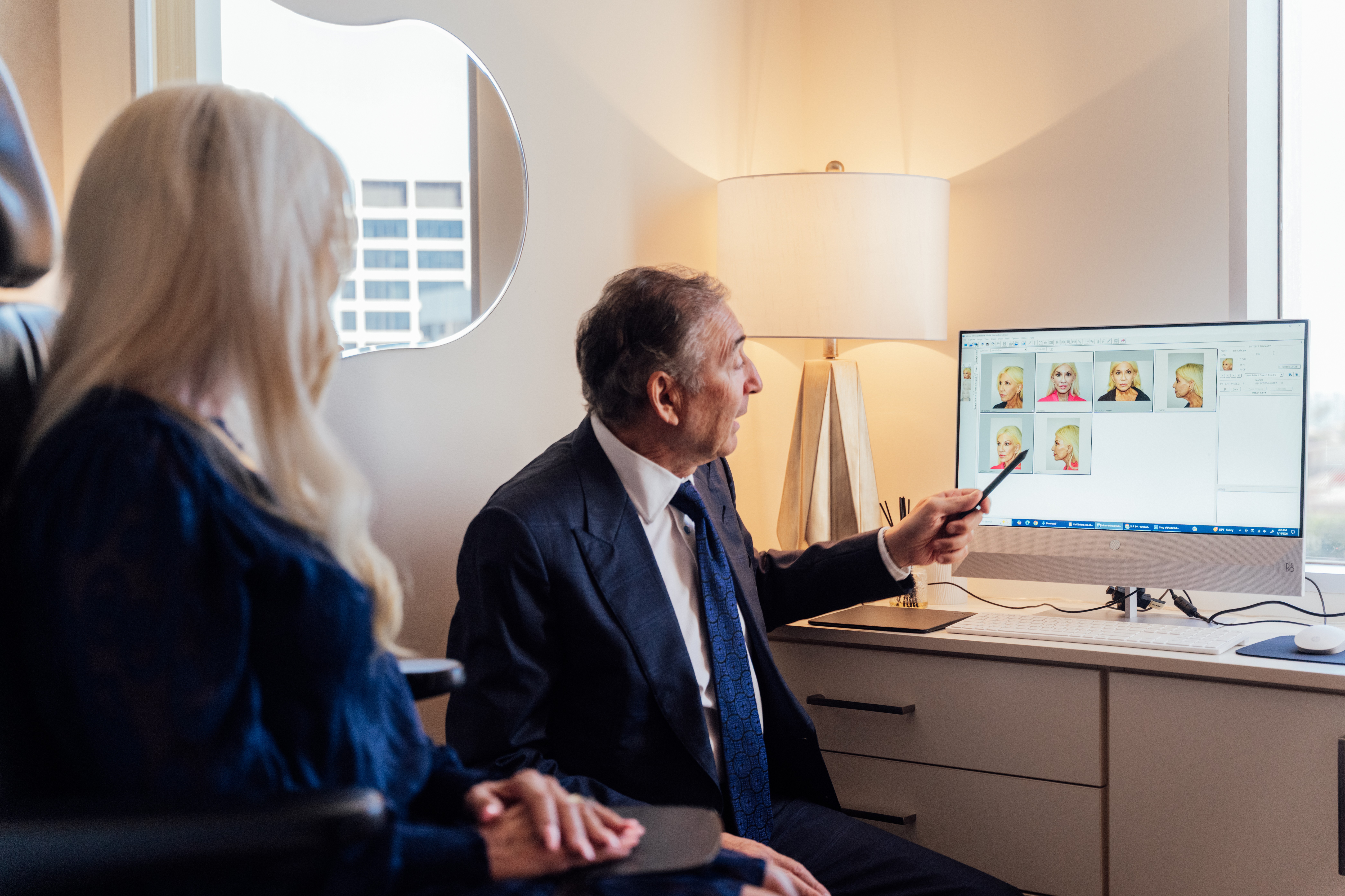 Two people in a room looking at a computer screen with images, one person pointing at the monitor, window and lamp in background.