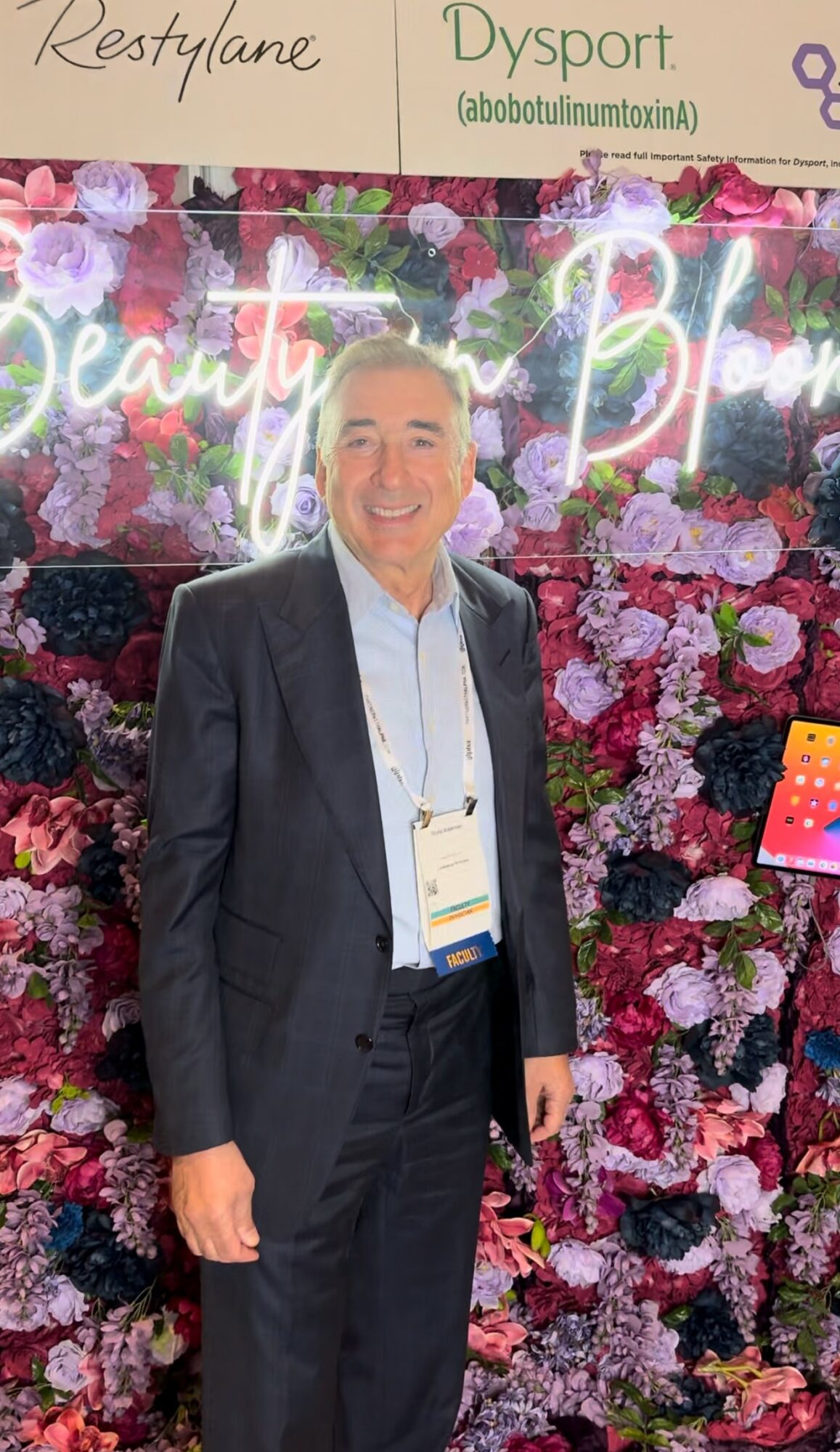 Man in suit standing in front of floral backdrop with neon sign reading 'Beauty'.