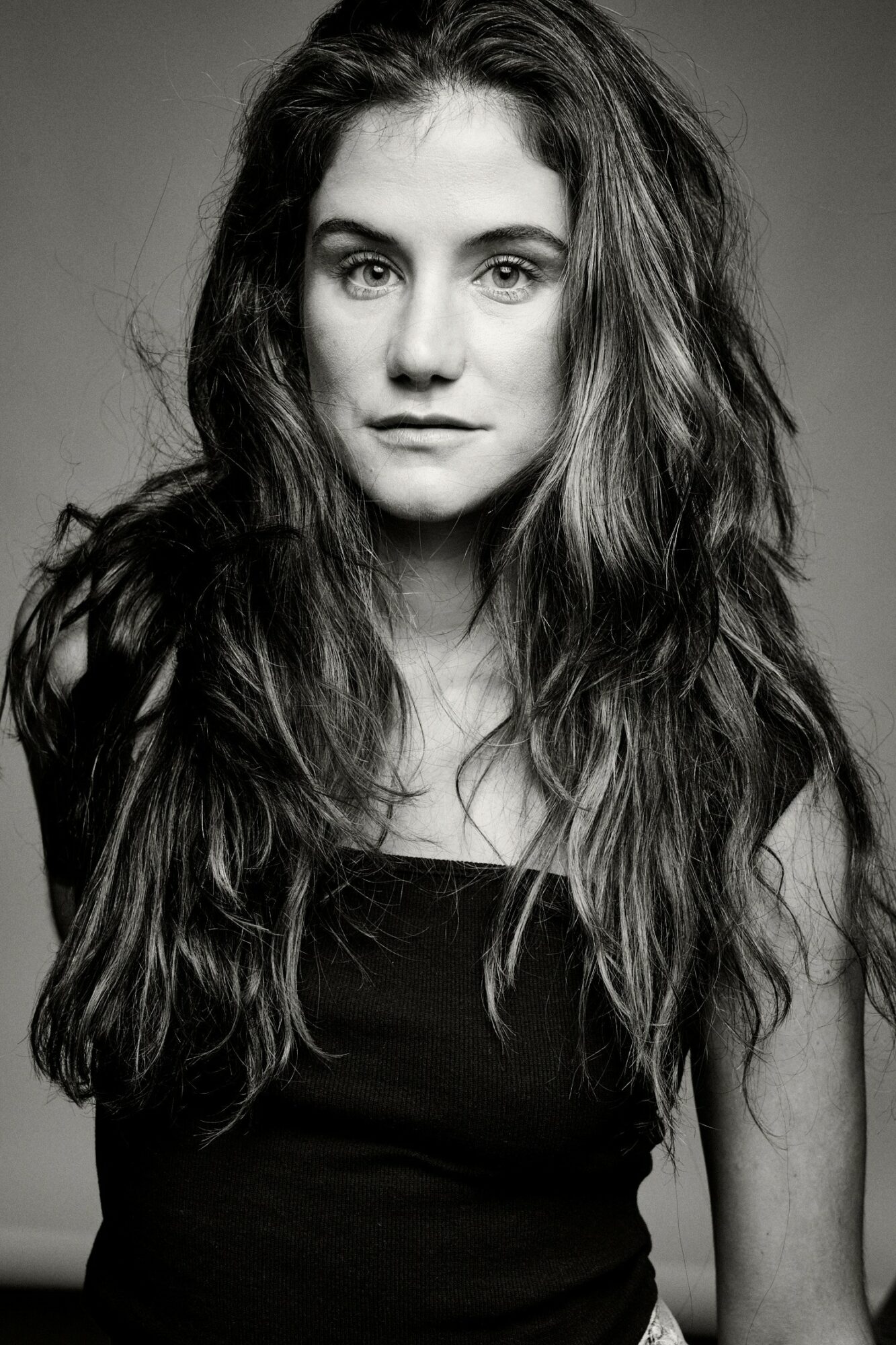 Young woman with long, wavy hair looking directly at the camera, wearing a dark top, black and white photo.