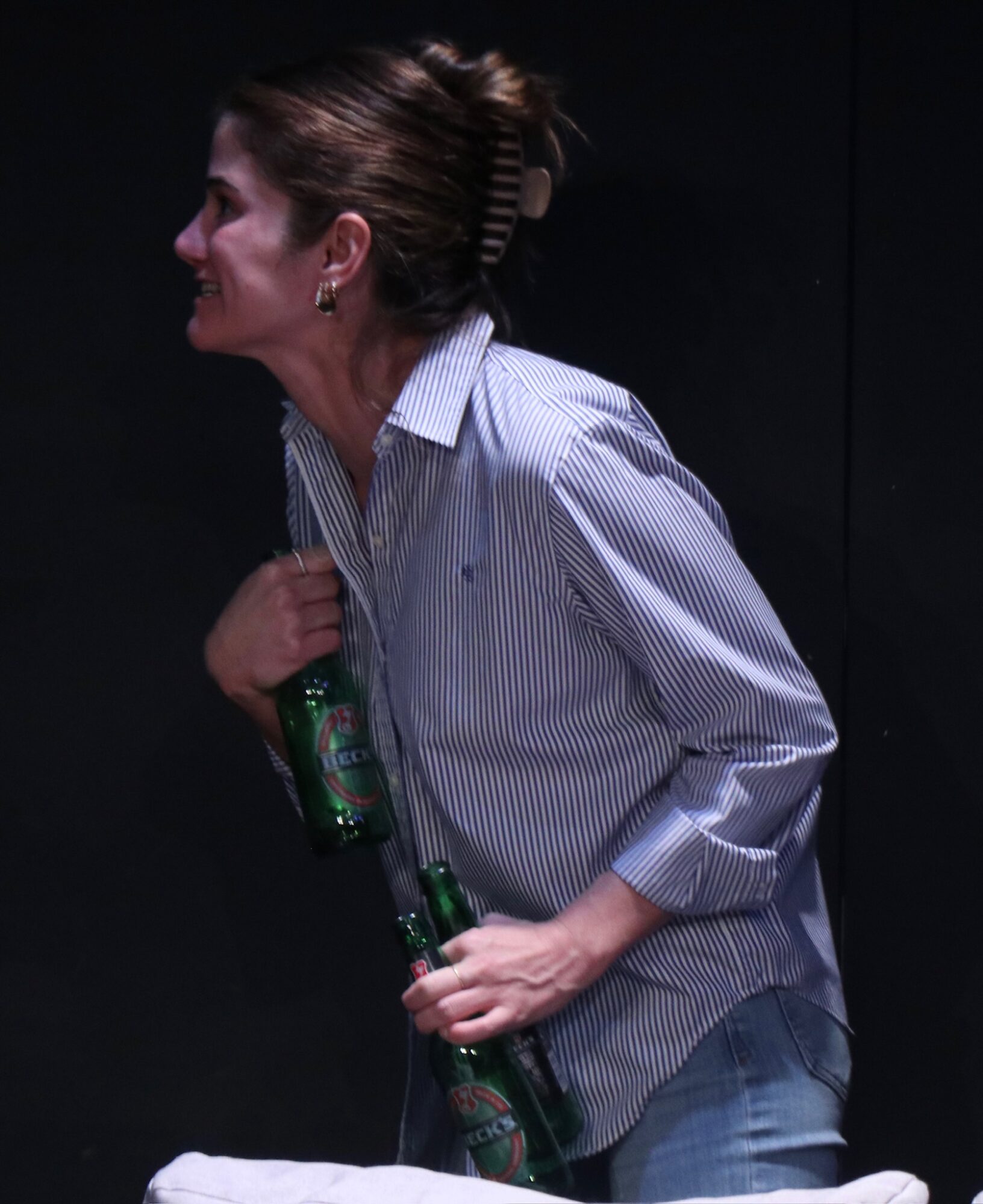 Woman with hair in a bun holding two green bottles, wearing a striped shirt, against a dark background.