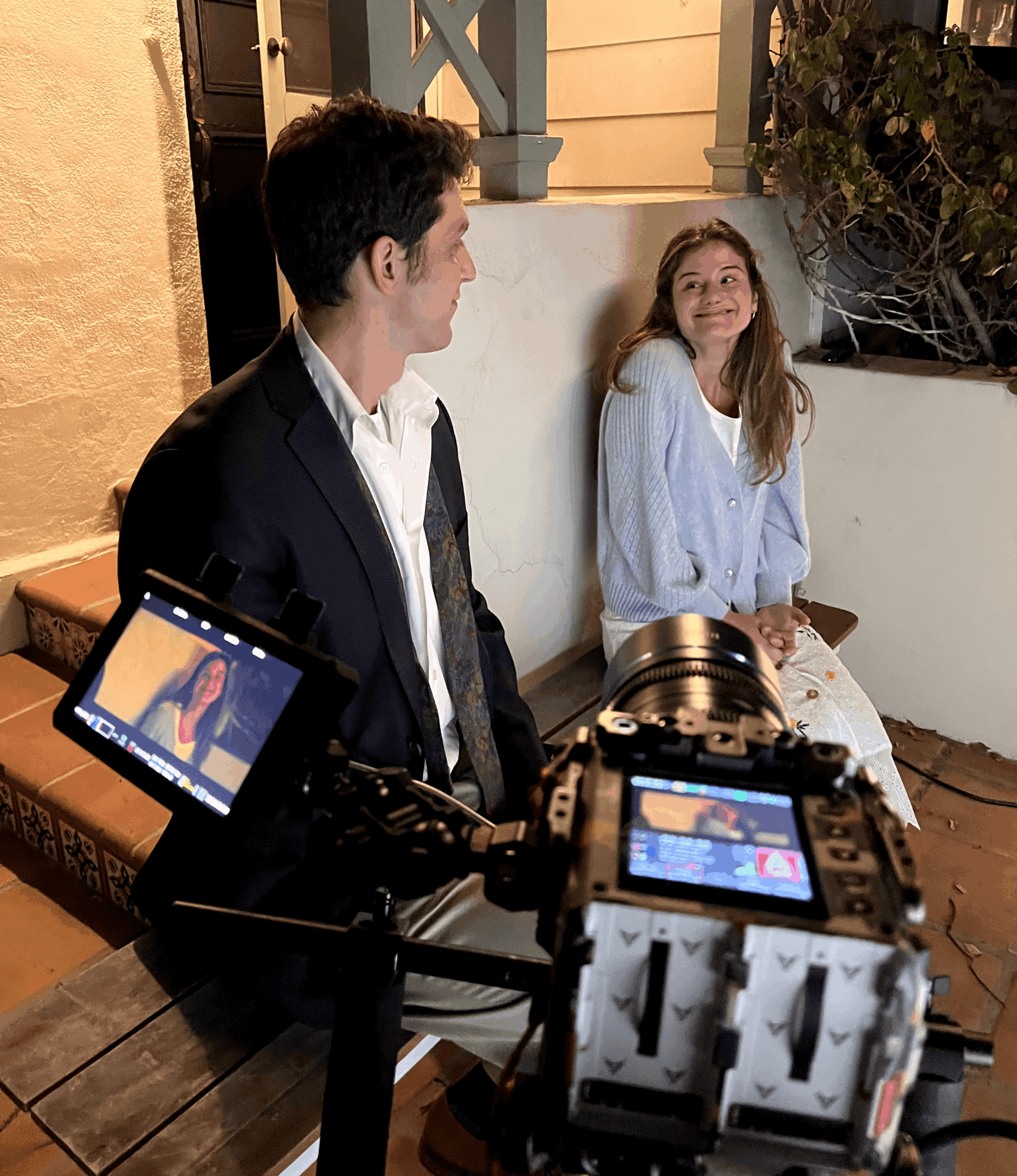 A young man and girl sit outdoors at night, smiling and looking at each other, with camera equipment in foreground.