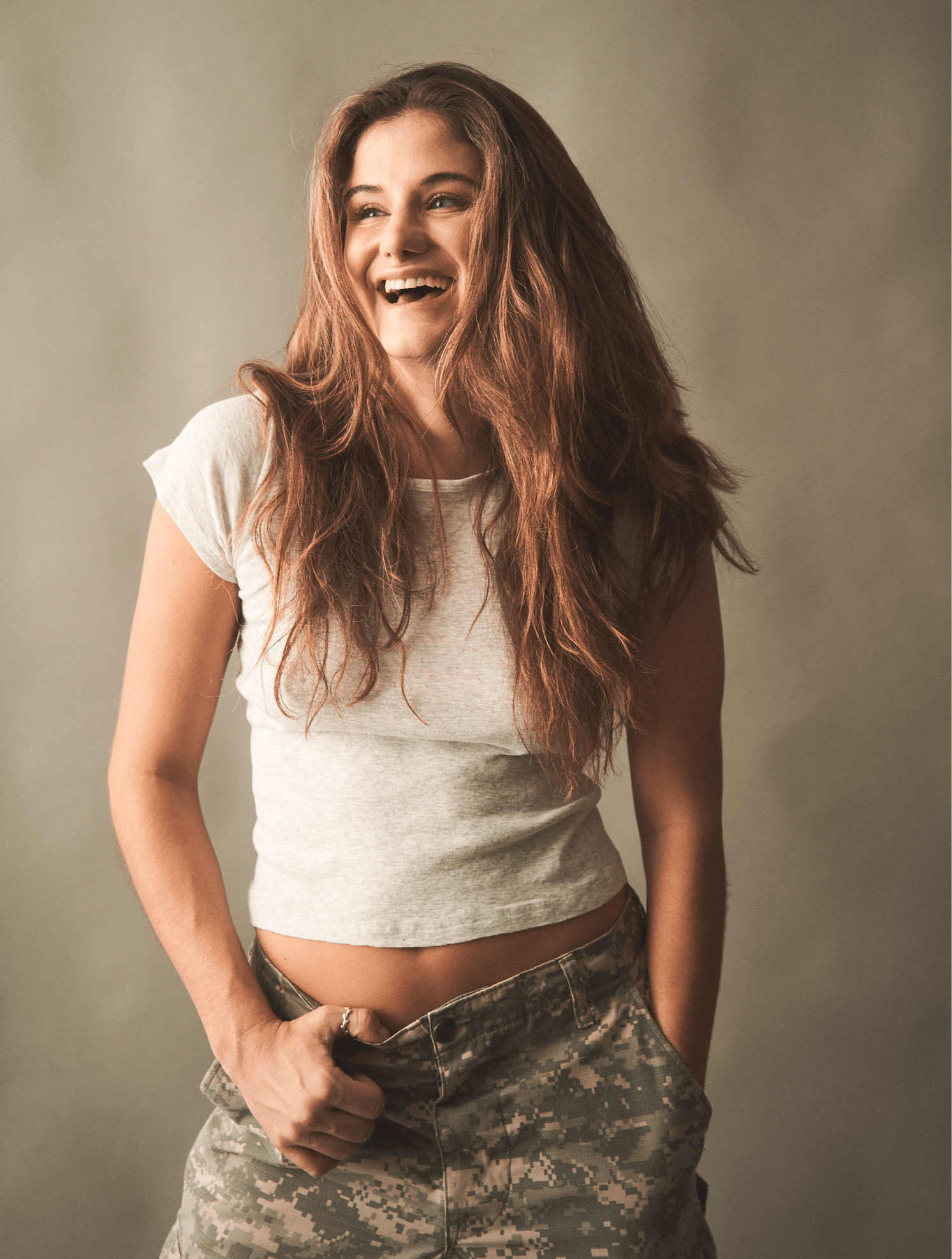 Young woman with long, wavy hair smiling, wearing a white t-shirt and camouflage pants, standing against a neutral background.