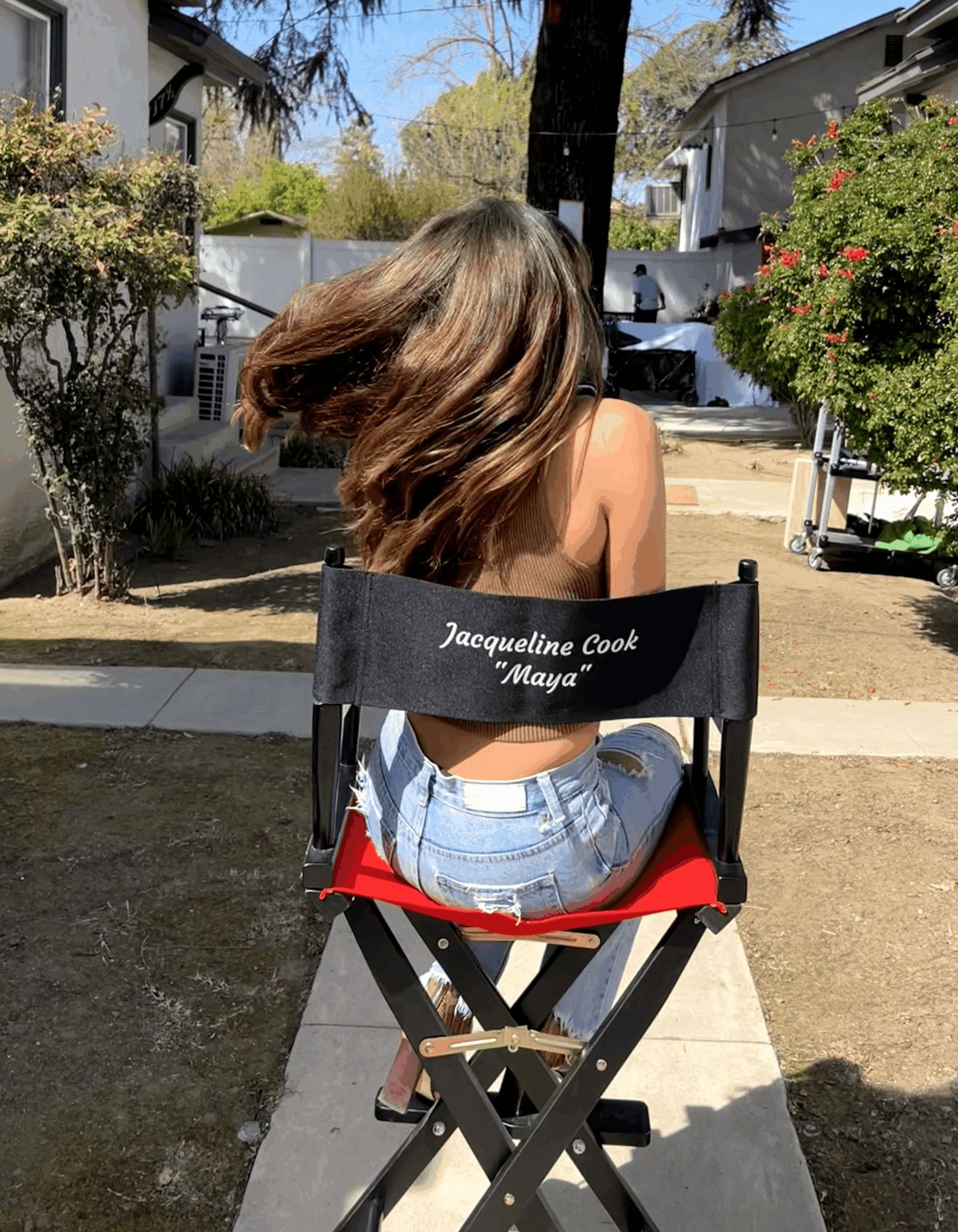 Person with long hair sitting on a red and black outdoor chair in a backyard, facing away, with trees and houses in background.