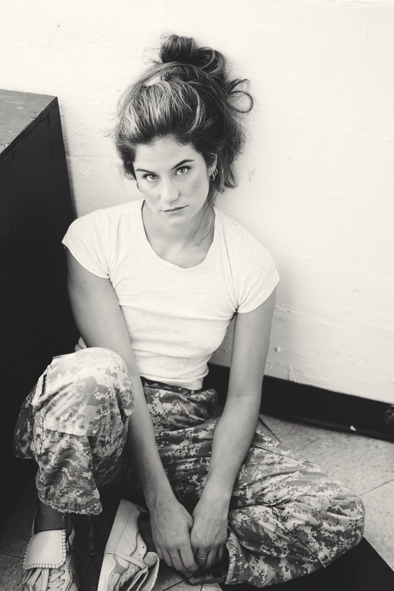 Young woman with messy hair sitting on the floor, wearing a white t-shirt and patterned pants, looking at the camera.