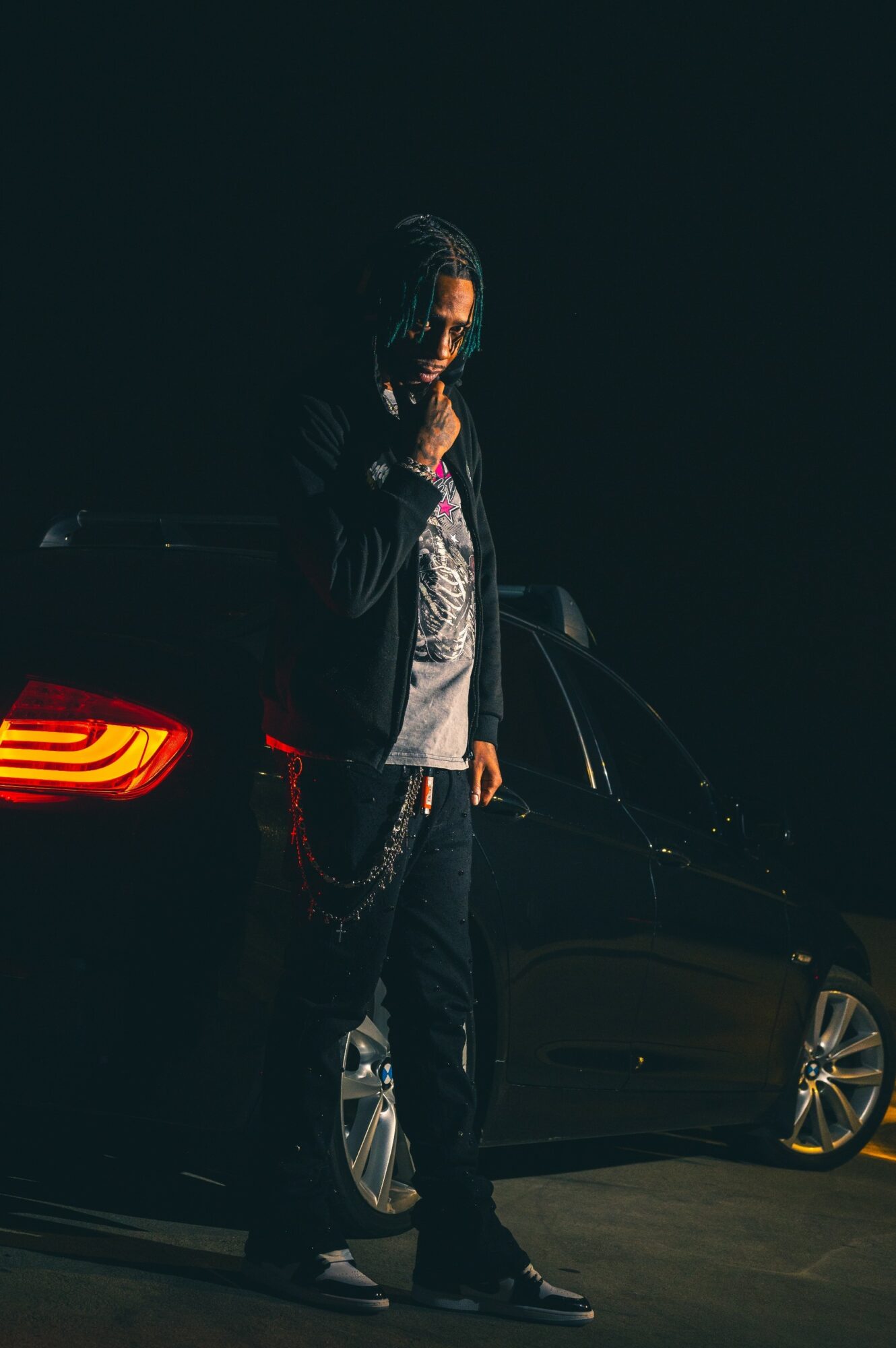 Person standing next to a black car in a dark setting, wearing glasses and casual clothing.