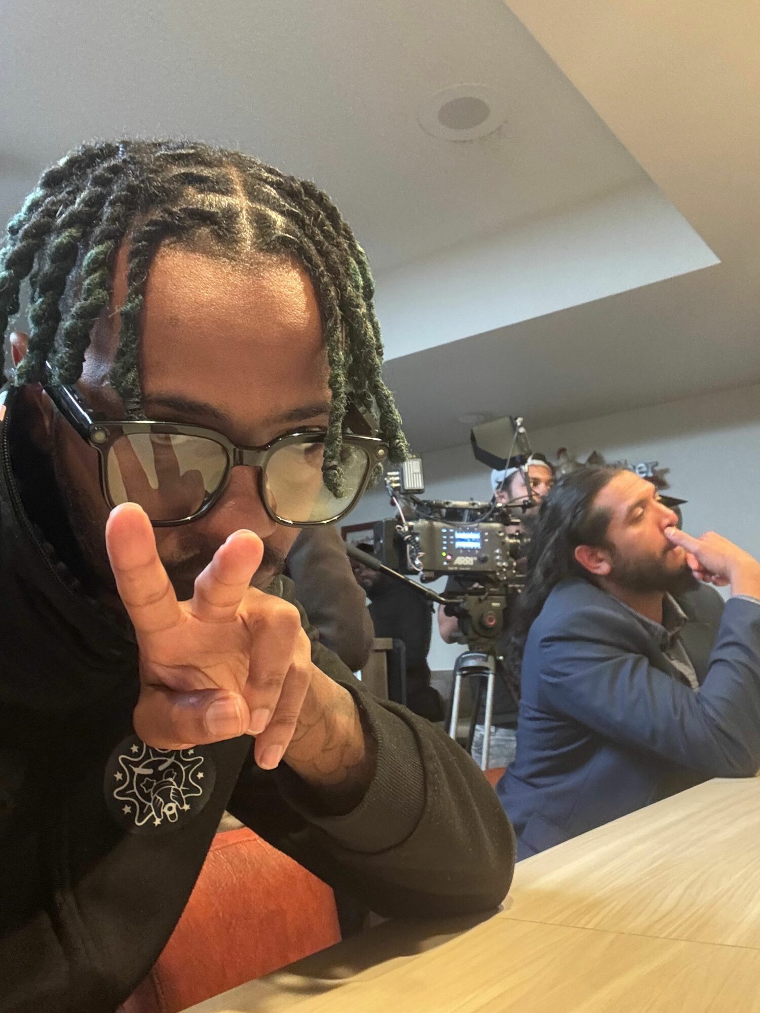 Person with braided hair and sunglasses making a peace sign, sitting on the floor, with a person in the background using camera equipment.