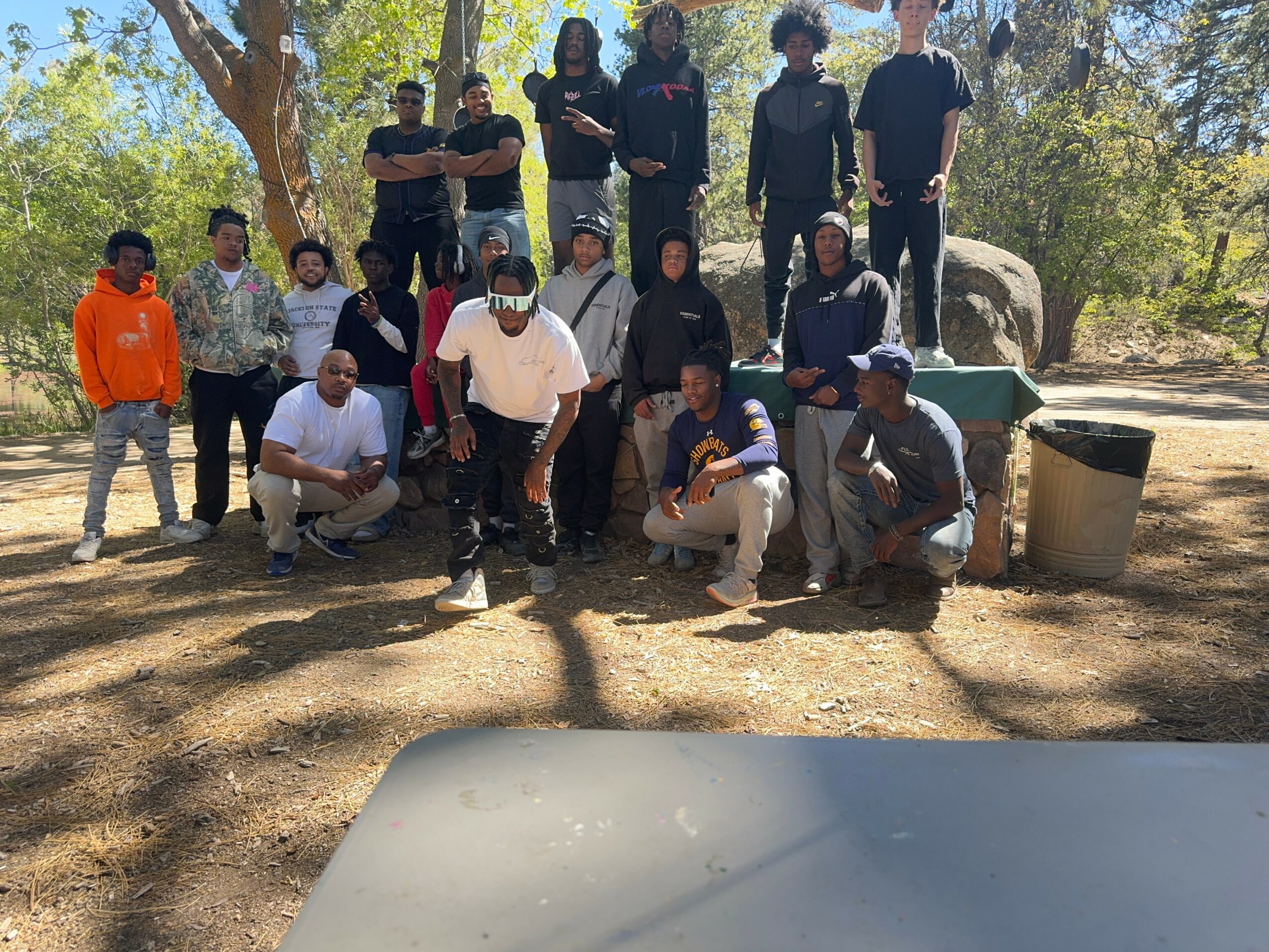 Group of people outdoors, some standing and some kneeling, with trees and rocks in the background.