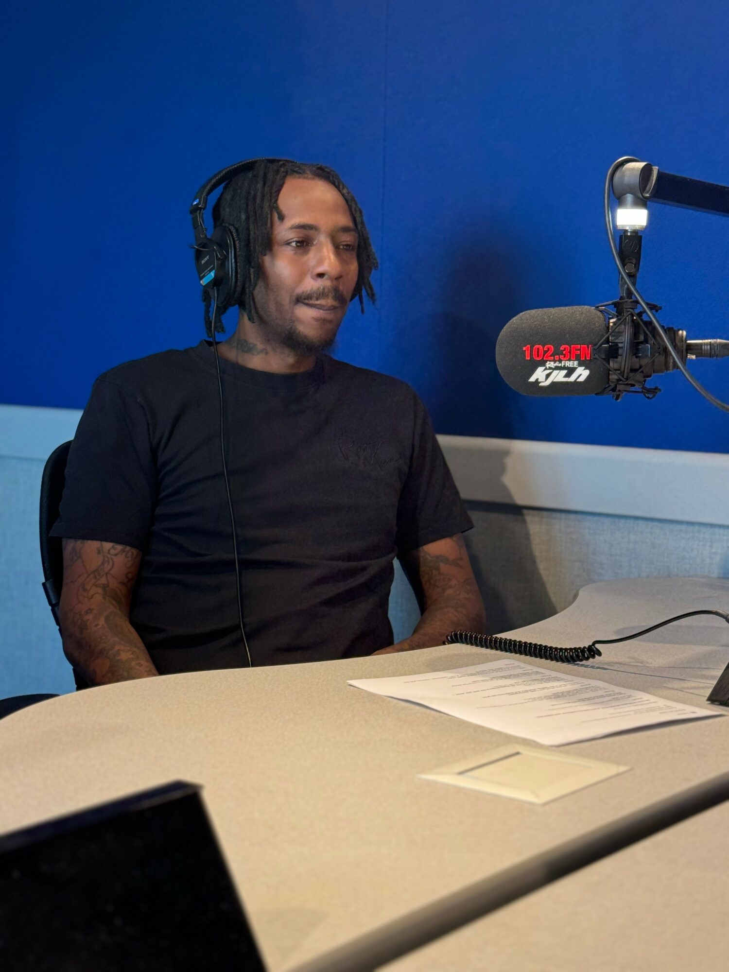 Person with headphones sitting at a table with papers, in a radio studio with a blue wall.