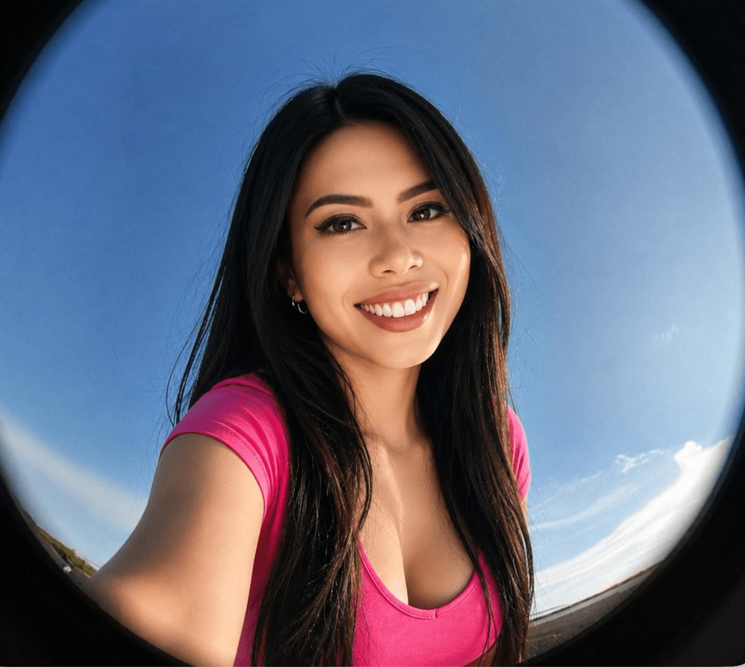 Smiling woman with long dark hair wearing a pink shirt against a blue sky background.