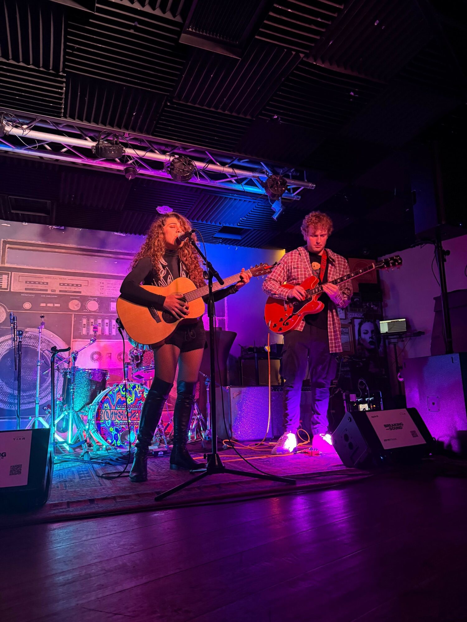 Two women perform with guitars on a stage illuminated by purple and blue lights, with sound equipment around them.