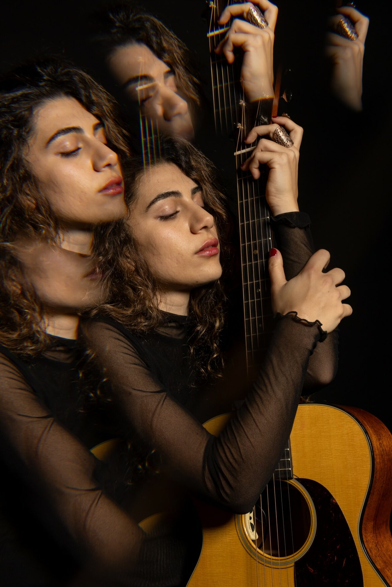 Three women with closed eyes holding a guitar, with reflections in a mirror behind them, dark background, soft lighting.