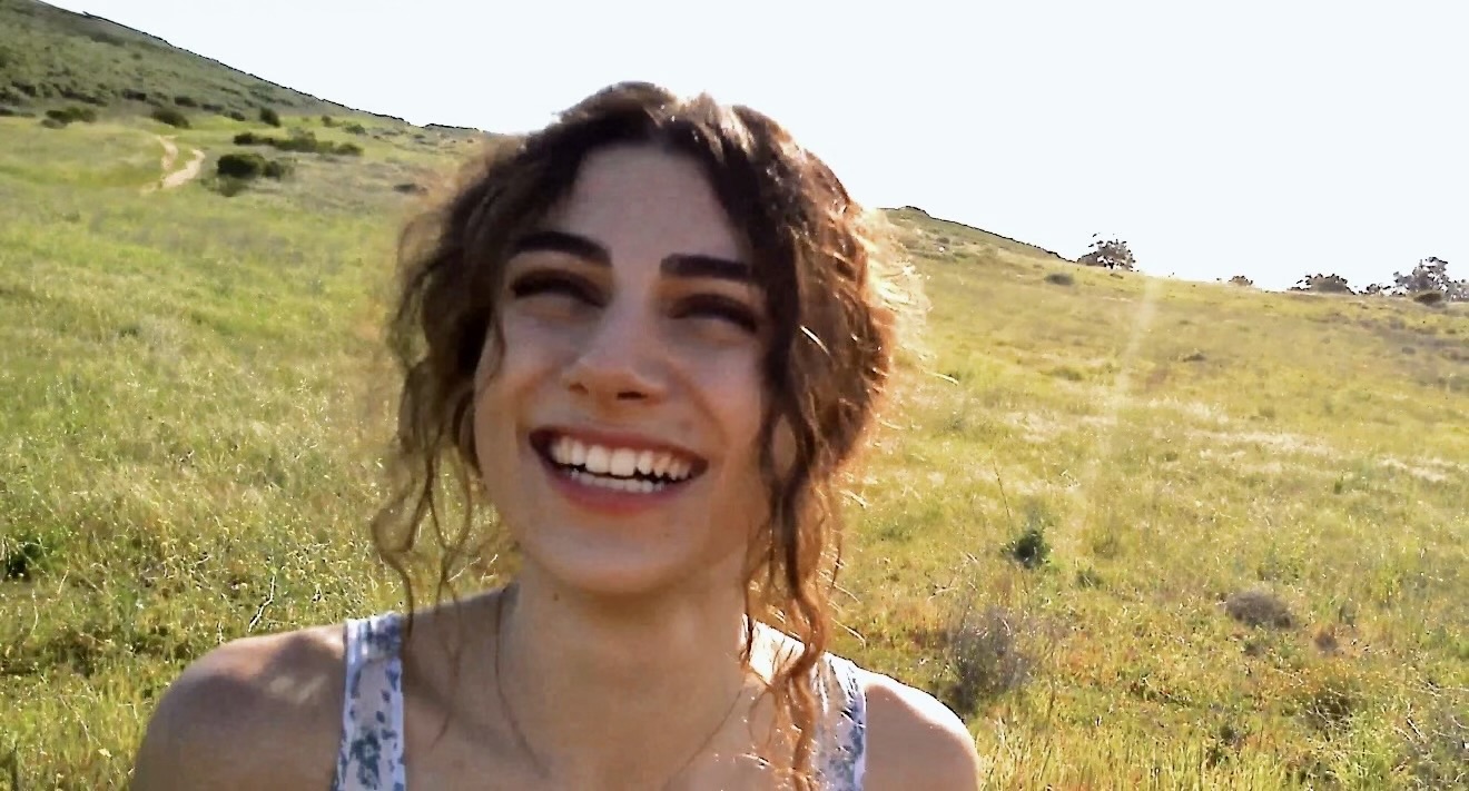 Young woman with curly hair smiling outdoors in a grassy field under bright sky
