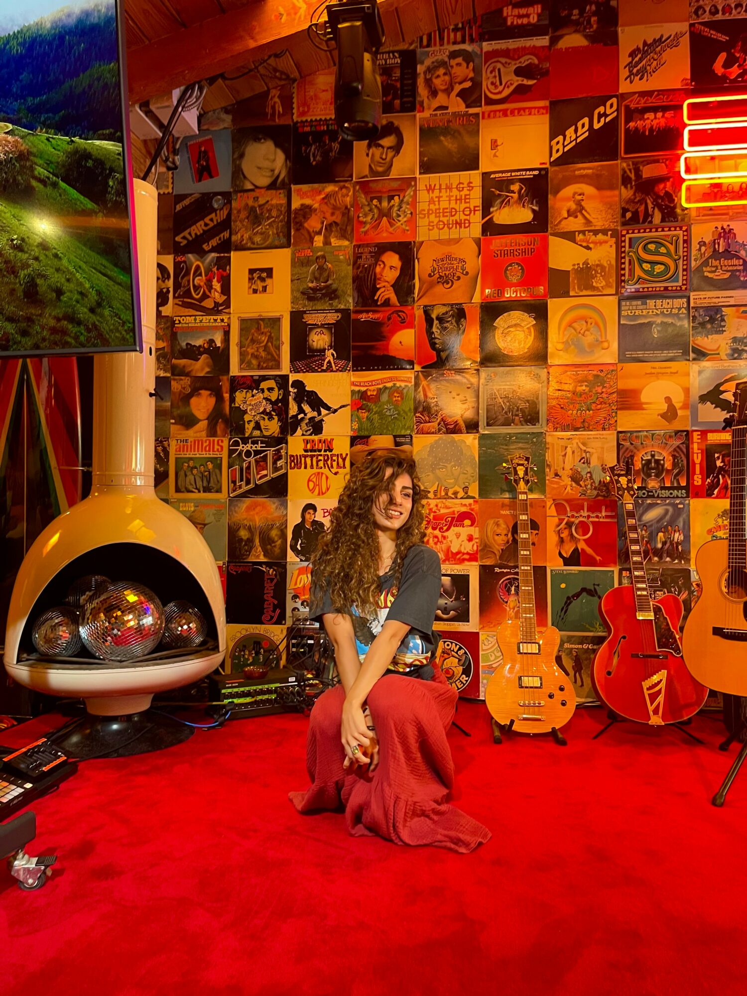 Person sitting on red carpet in front of a wall covered with colorful album covers, with guitars nearby, in a music-themed room.