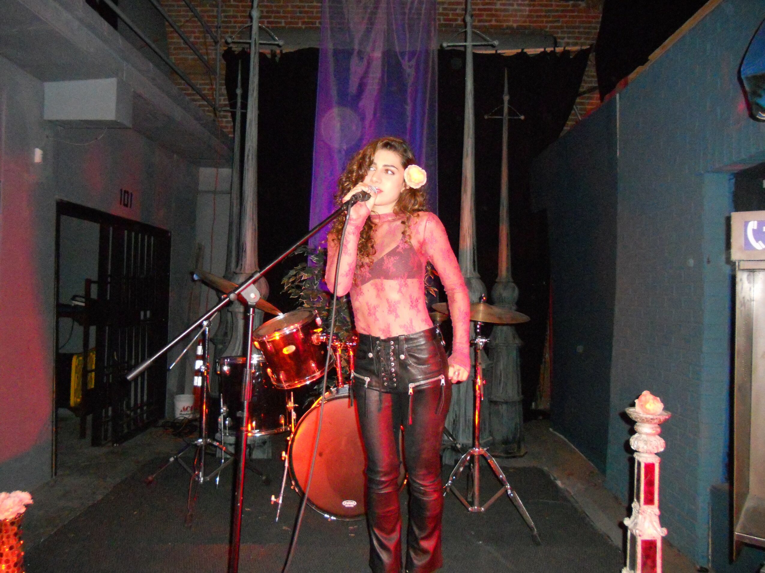 Female singer with curly hair performs on stage with a drum set behind her, wearing a pink top and black pants.