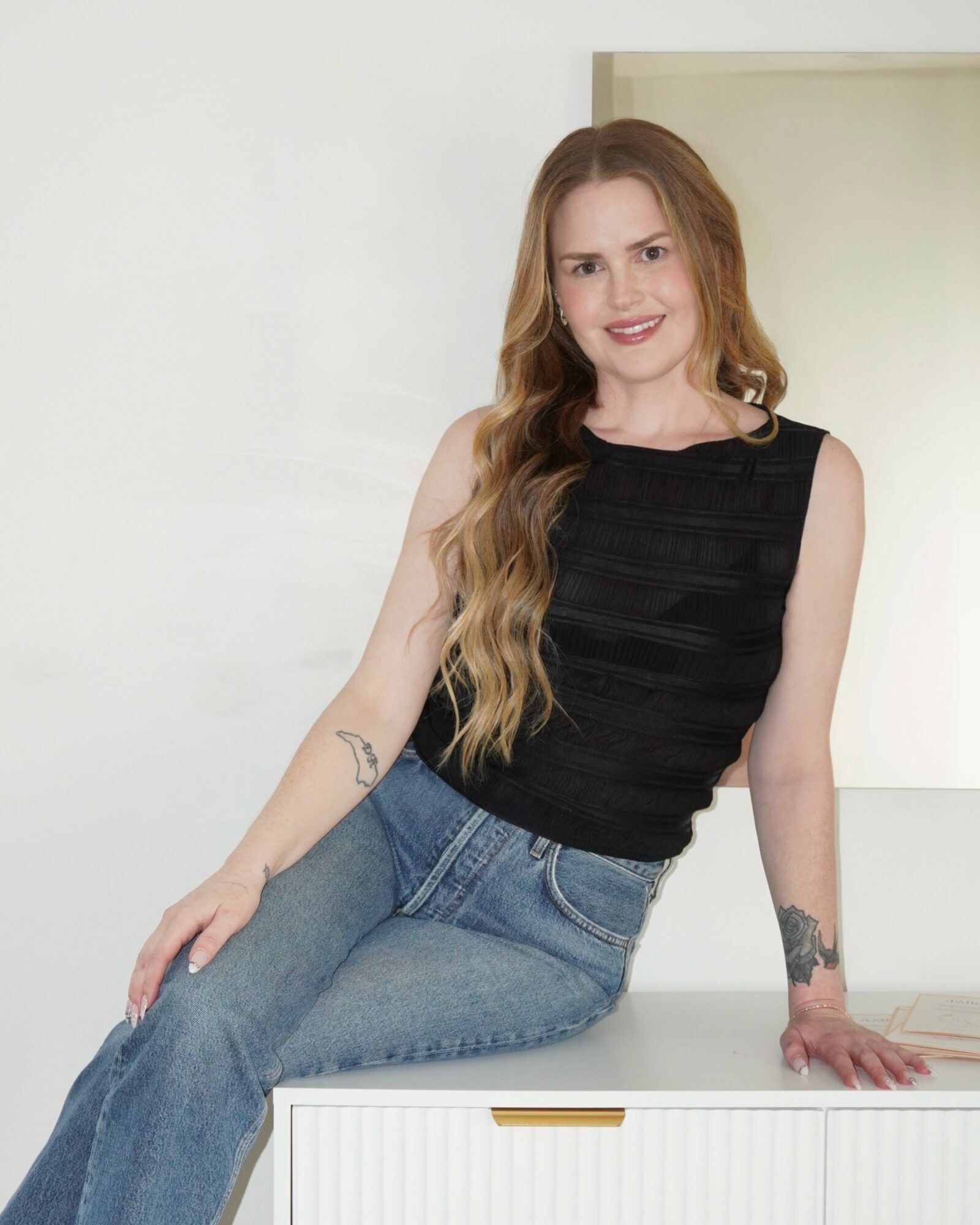 Young woman with long wavy hair, wearing a black sleeveless top and blue jeans, sitting on a white surface, smiling.