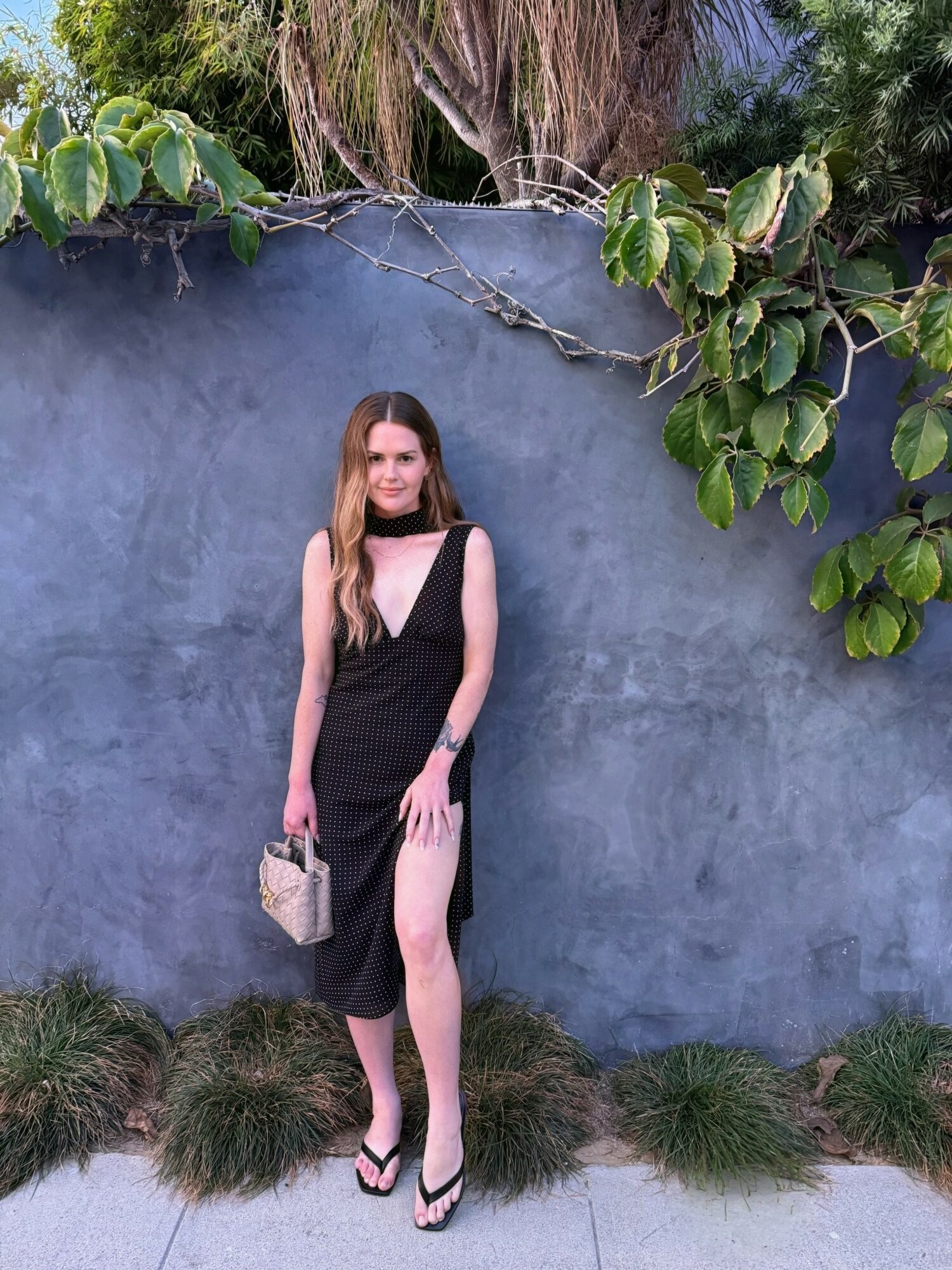 Young woman in black dress and heels standing against a blue wall with green foliage, holding a small bag.