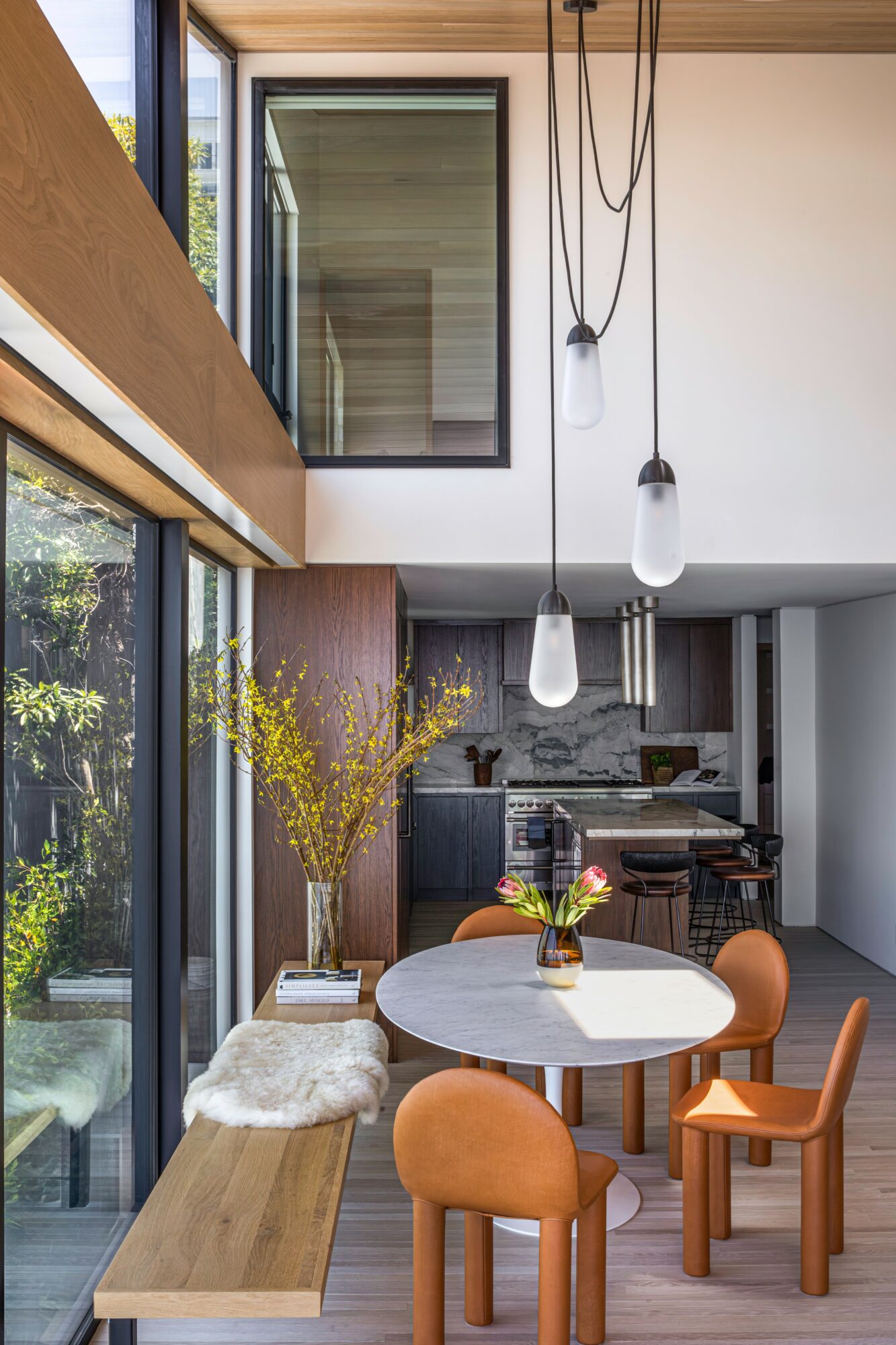 Modern dining area with round table, four orange chairs, large window, and hanging light bulbs, with a vase of flowers.