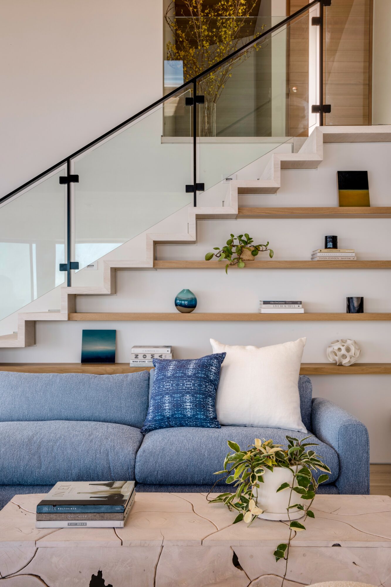 Living room with a blue sofa, white pillows, and a wooden table with a plant, staircase with glass railing in background.