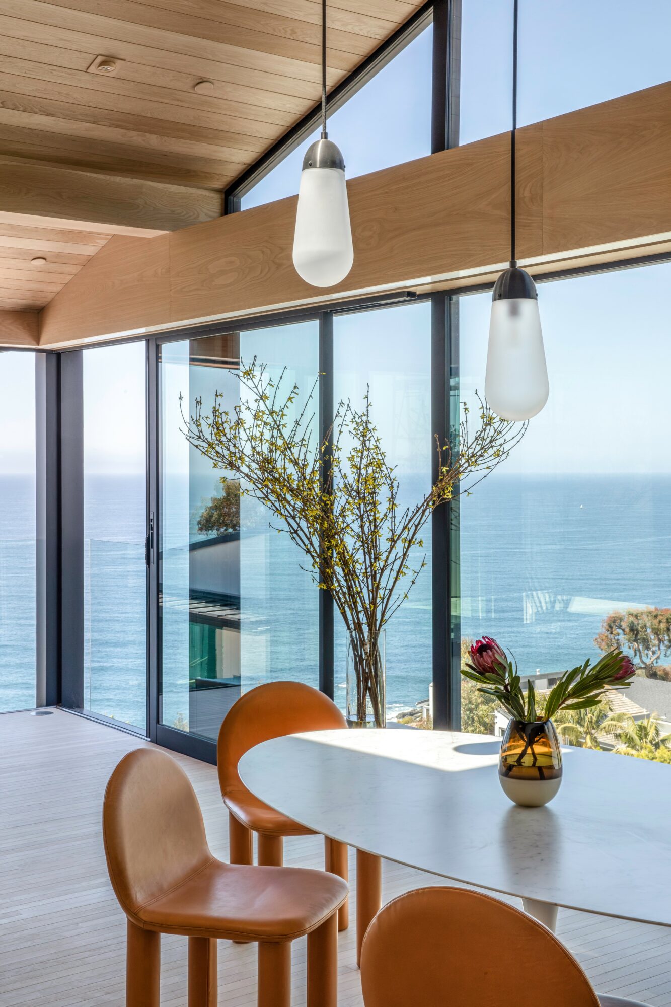 Interior with large windows, a round table with a vase and flowers, and hanging pendant lights, overlooking the ocean.