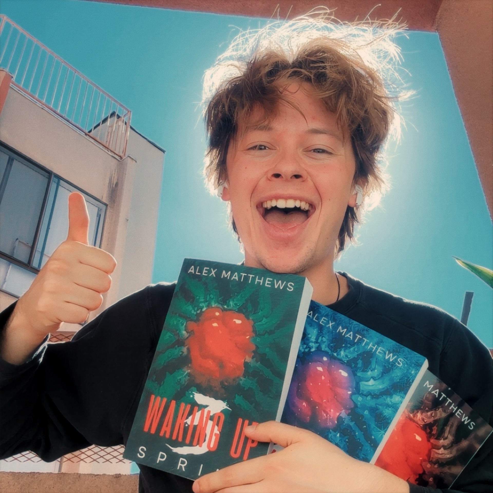 Young person with messy hair smiling and giving a thumbs-up, holding three books titled 'Waiting Up' by Alex Matthews.