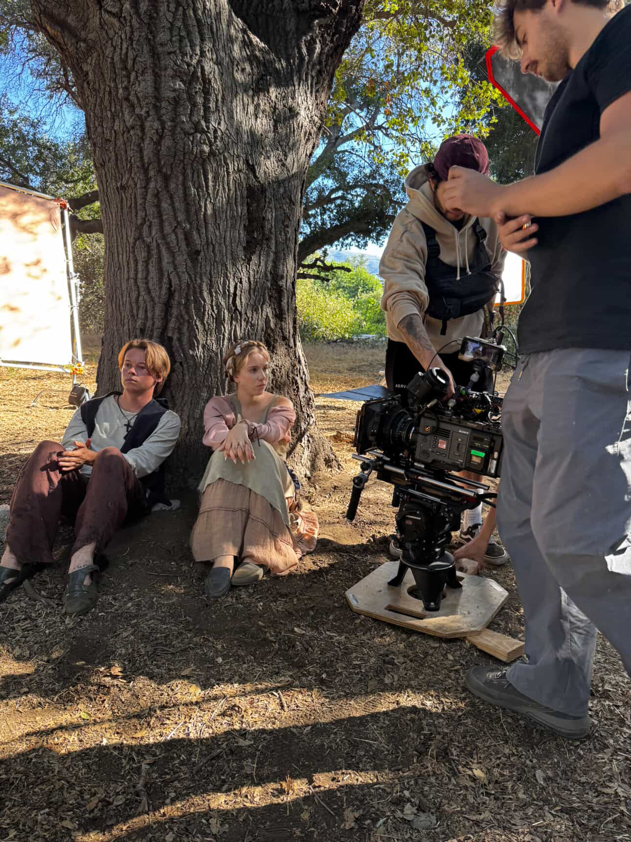 Two children sit under a large tree, while two adults operate a camera nearby outdoors.