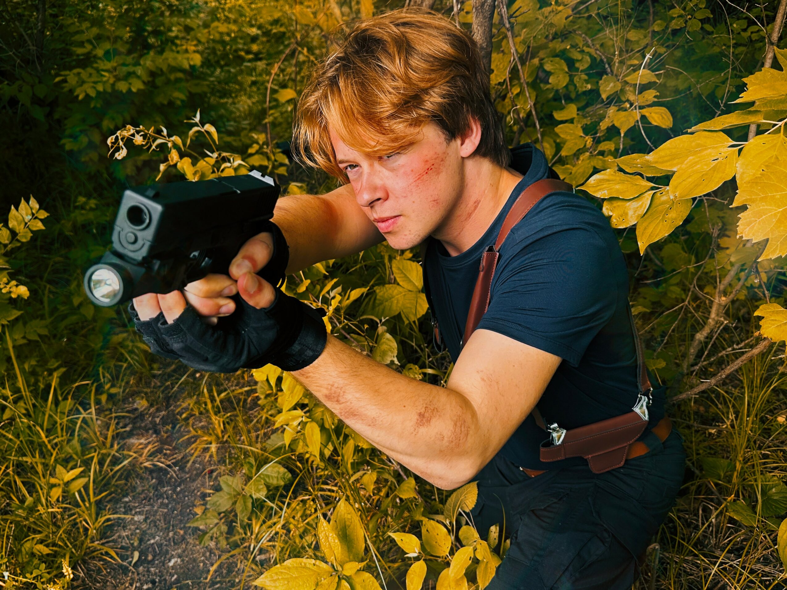 Young man with brown hair aiming a handgun outdoors among yellow and green foliage.