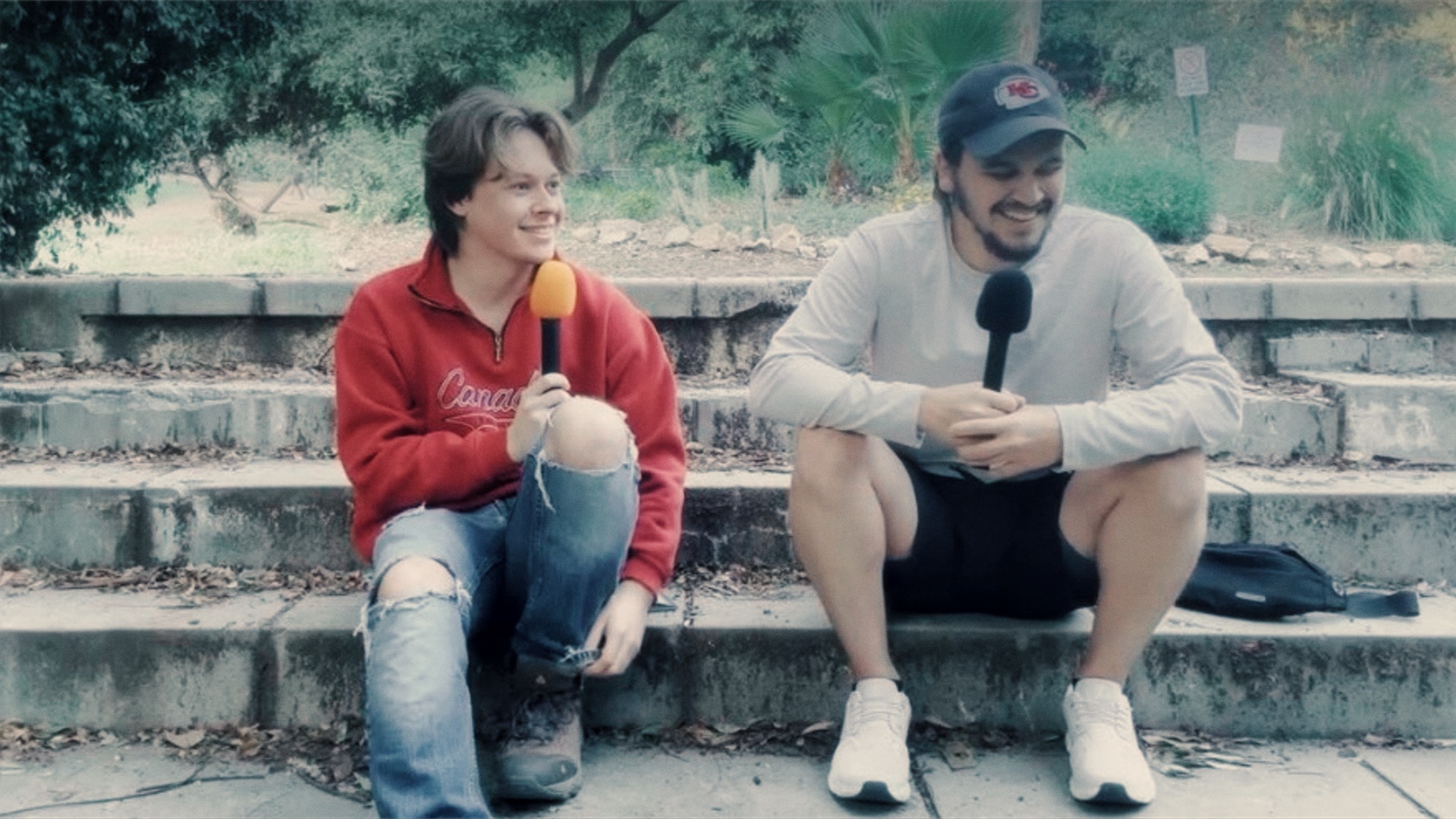 Two people sitting on outdoor steps, one holding a microphone, smiling, with trees in the background.