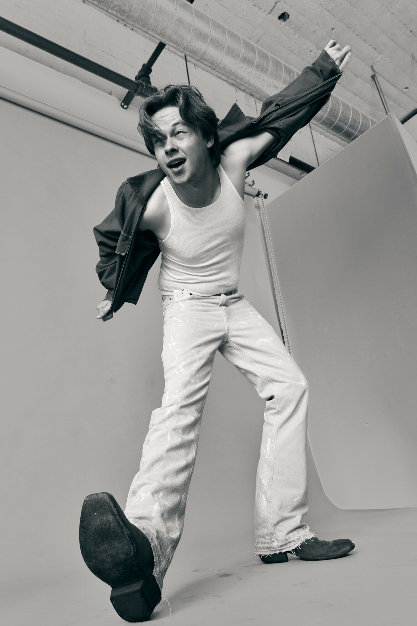 Person with open mouth, wearing a jacket and white pants, standing with one foot on the ground and the other raised, in a studio.
