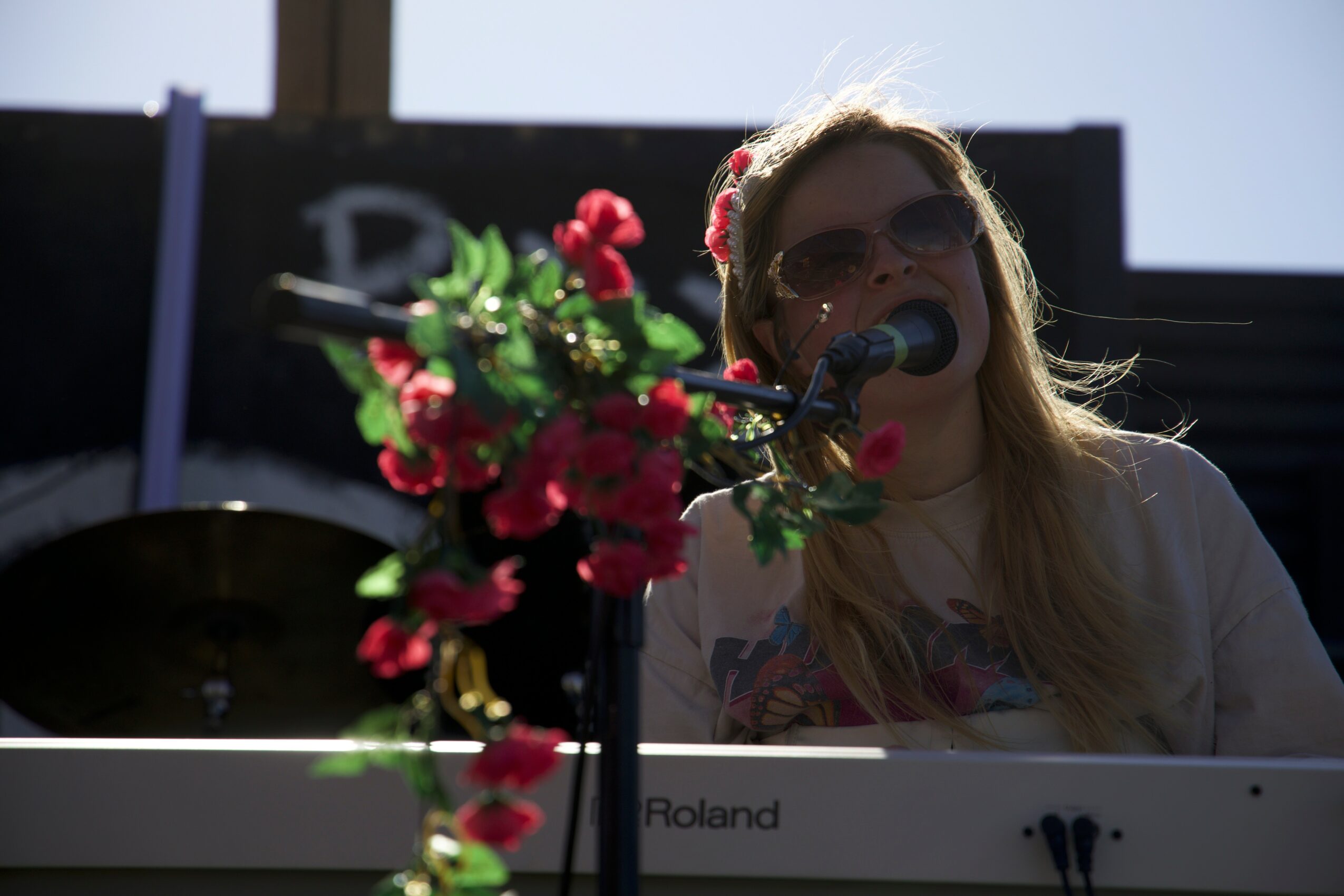 Person with long hair and sunglasses playing a keyboard, with a microphone and pink flowers in front, outdoors.