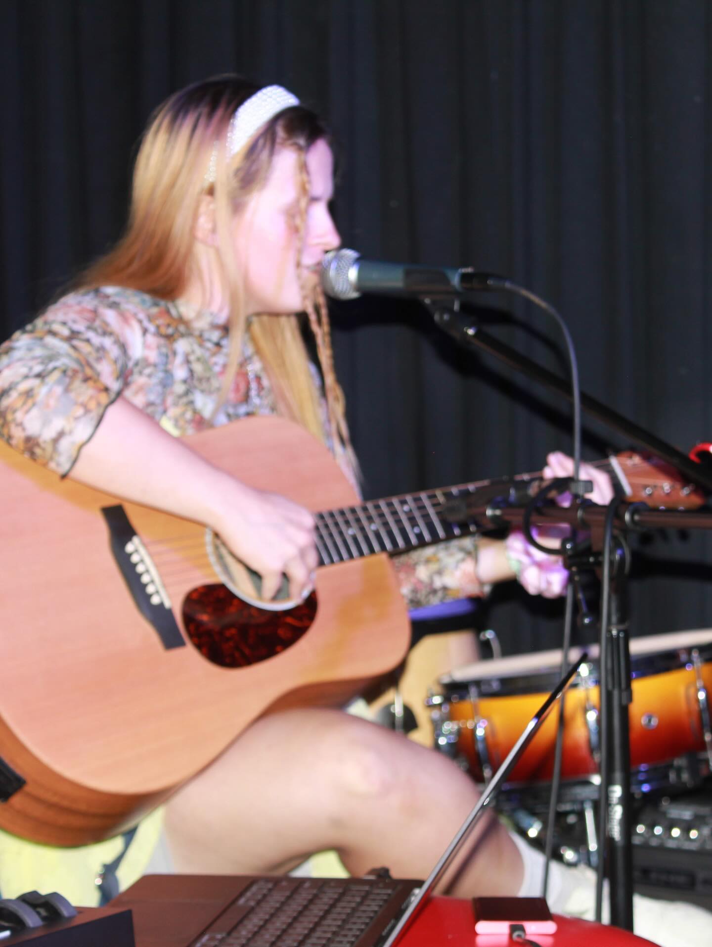 Young woman with long hair playing guitar and singing into a microphone on stage, with drums in background.