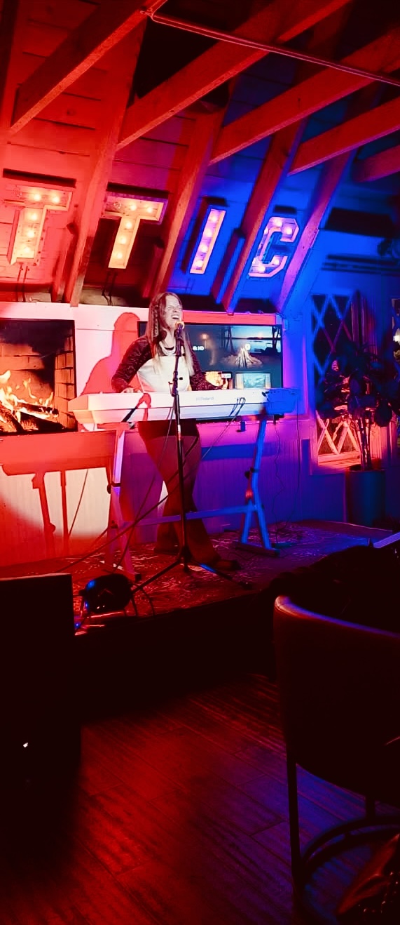 Person performing music on stage with red and blue lighting, in a venue with wooden beams and neon signs.
