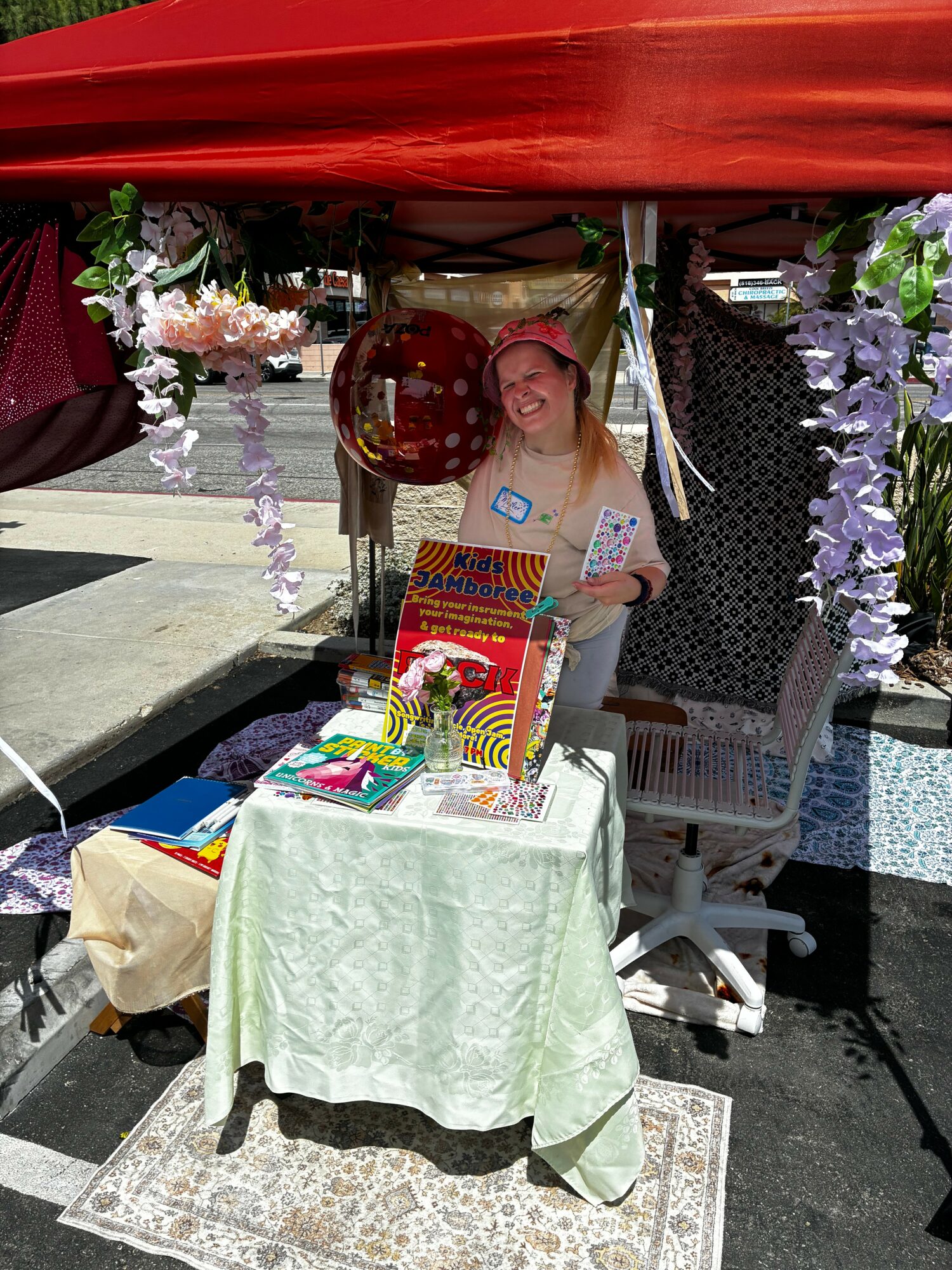 Woman standing behind a table with colorful books and decorations, under a red canopy, outdoors.