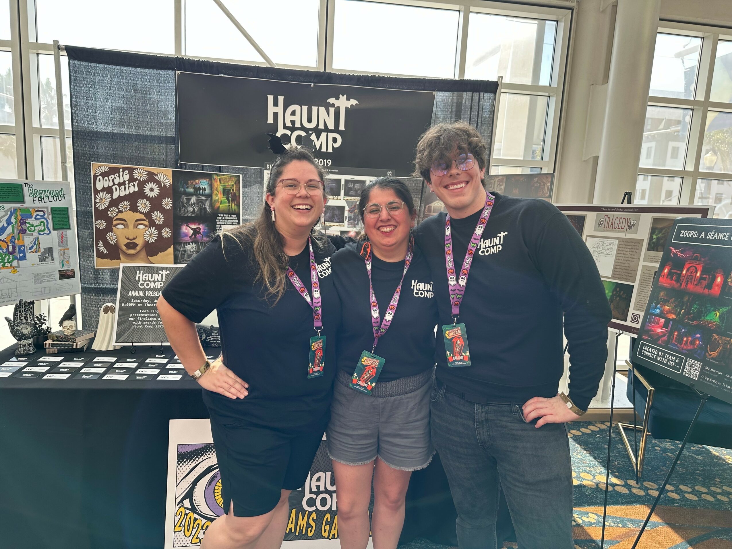 Three smiling people standing together at a booth with a 'Haunt Camp' sign behind them, inside a large room with windows.