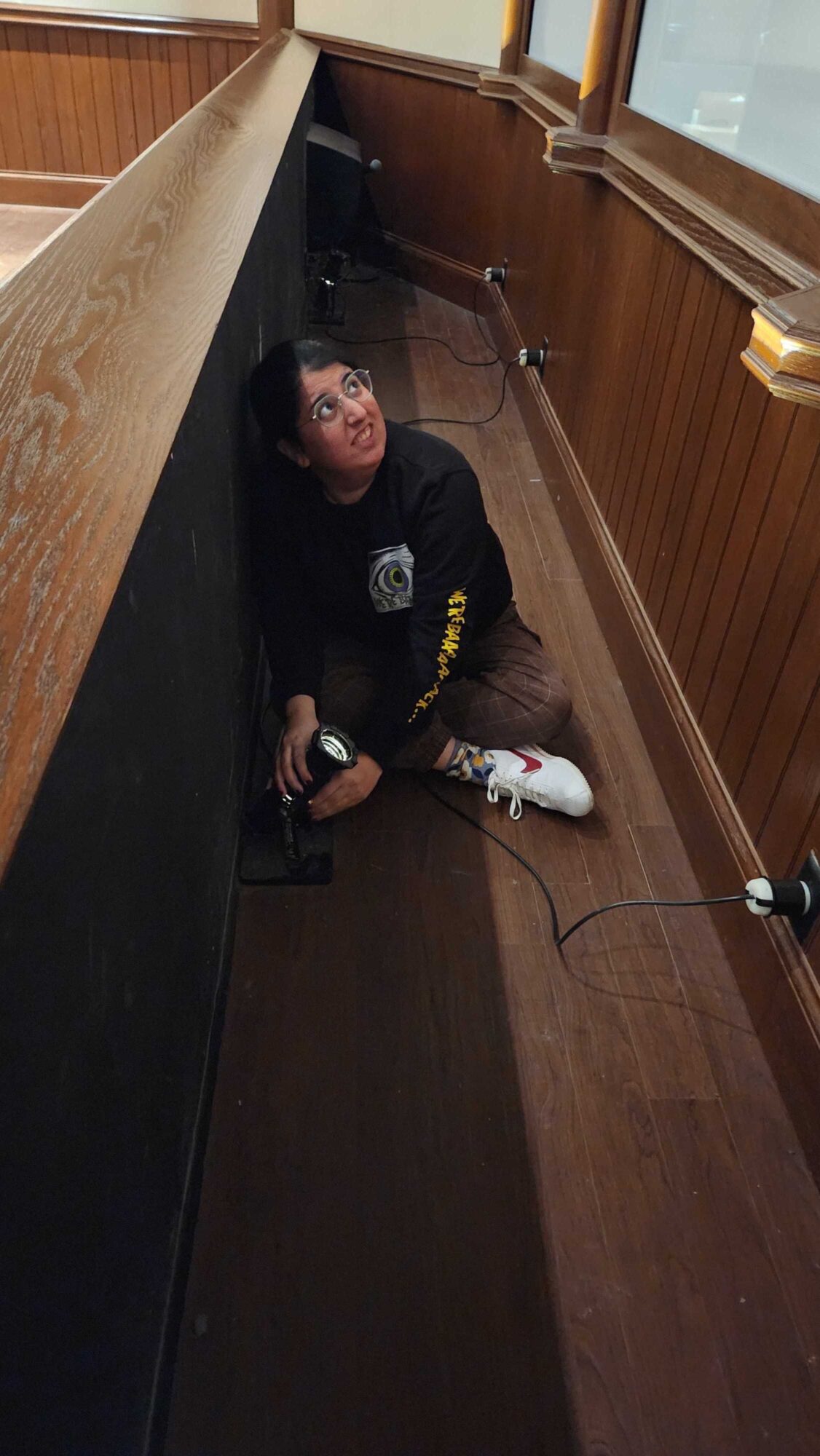 Person sitting on the floor in a narrow wooden-paneled room, looking up, with a phone in hand, and a cable on the floor.