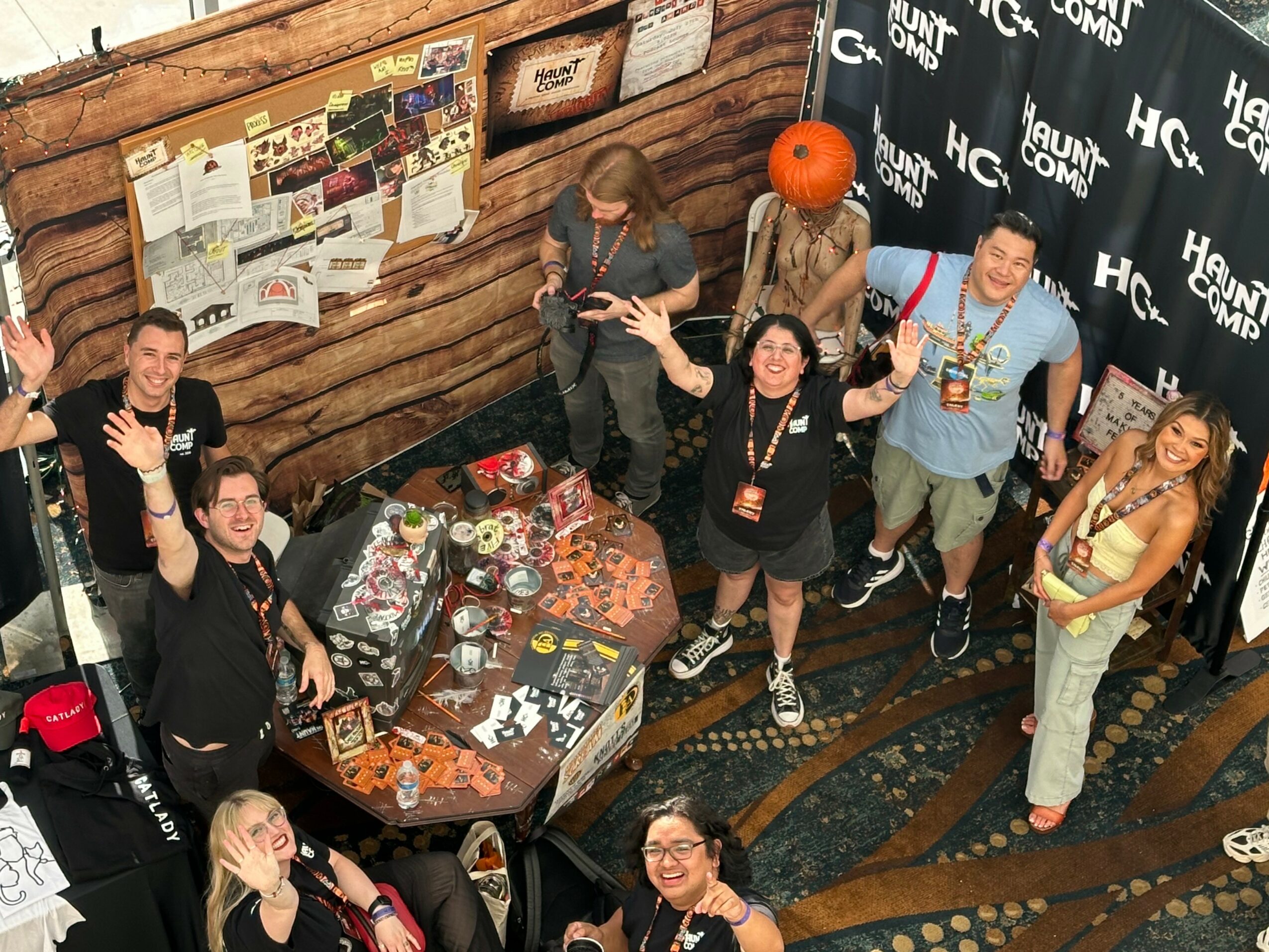 Group of seven people smiling and waving at camera in indoor event space with a table of items and a backdrop.