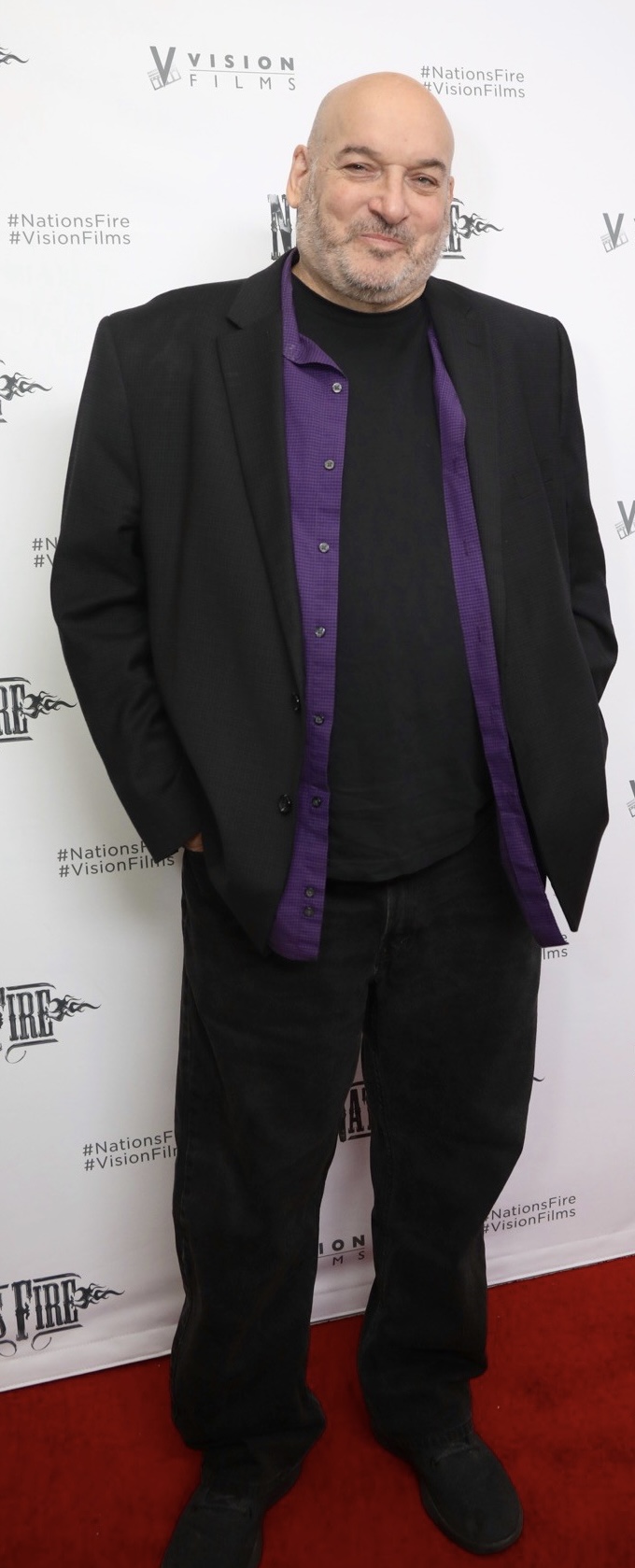 Bald man with a beard in a black suit and purple shirt, standing on a red carpet in front of a white backdrop.