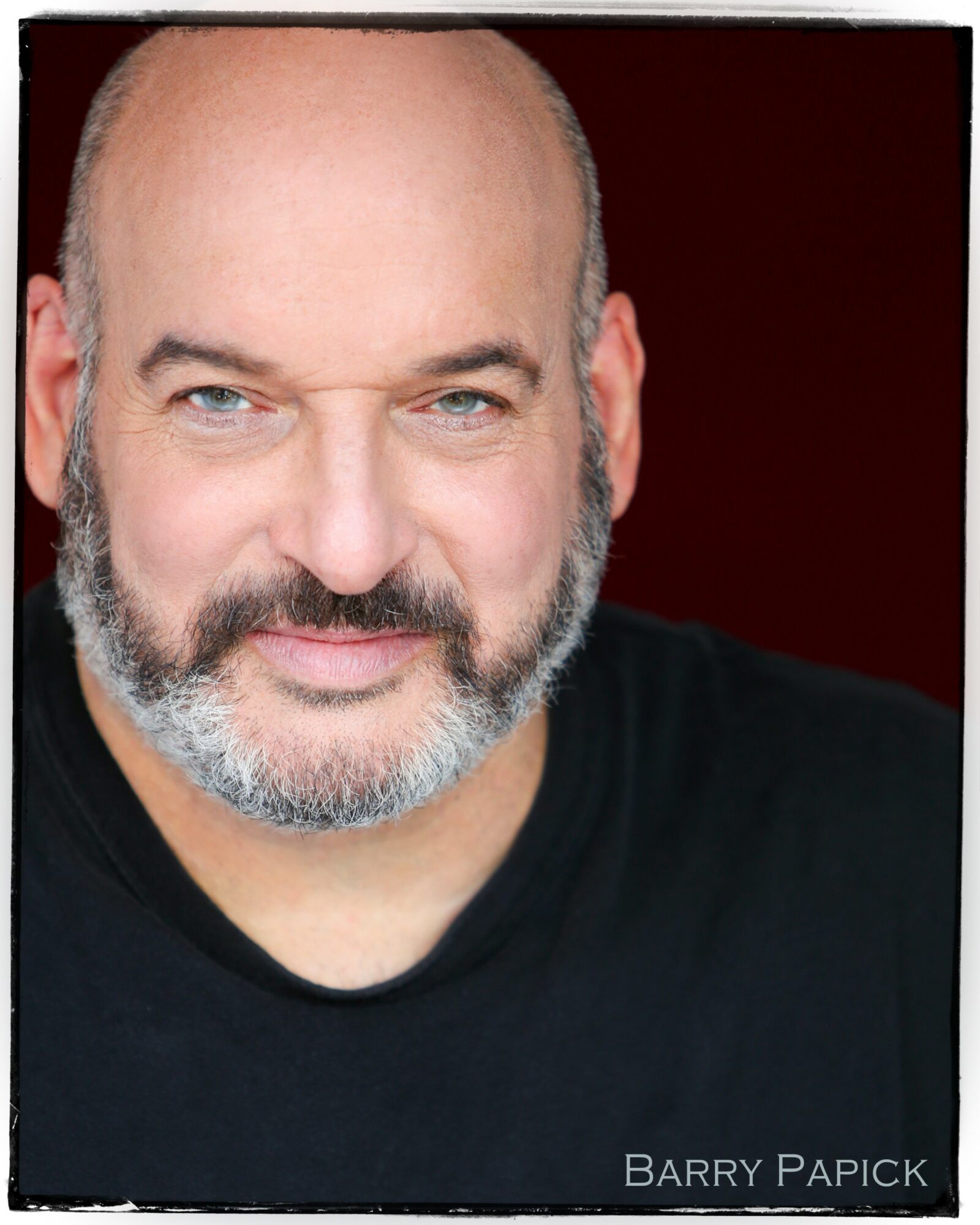 Close-up of a smiling man with a beard and blue eyes, wearing a black shirt, against a dark background.