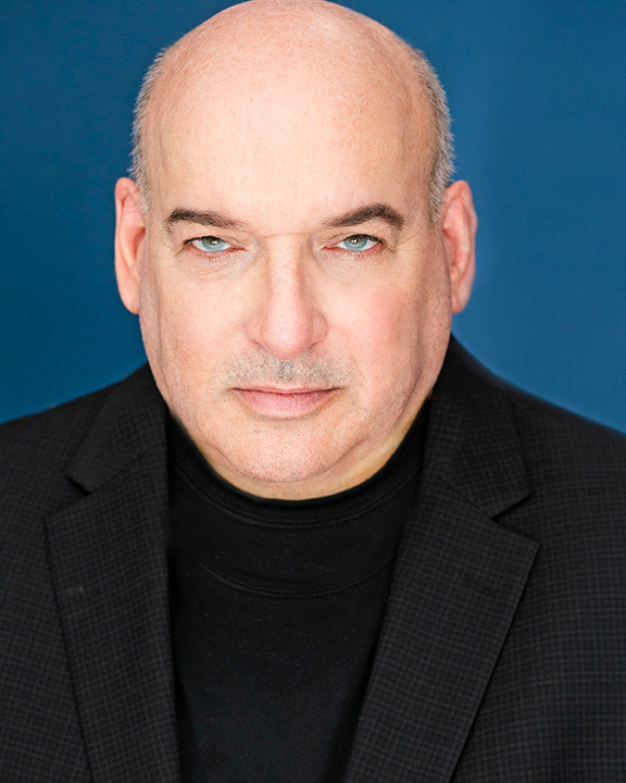 Portrait of a middle-aged man with a serious expression, bald head, wearing a black blazer and black shirt, against a blue background.