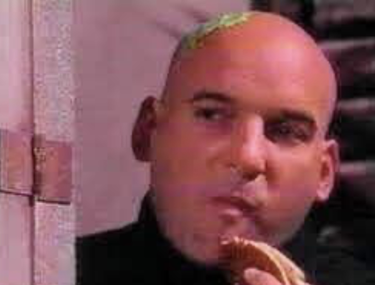 Bald man with a small patch of hair on top, holding a sandwich near his mouth, looking to the side.