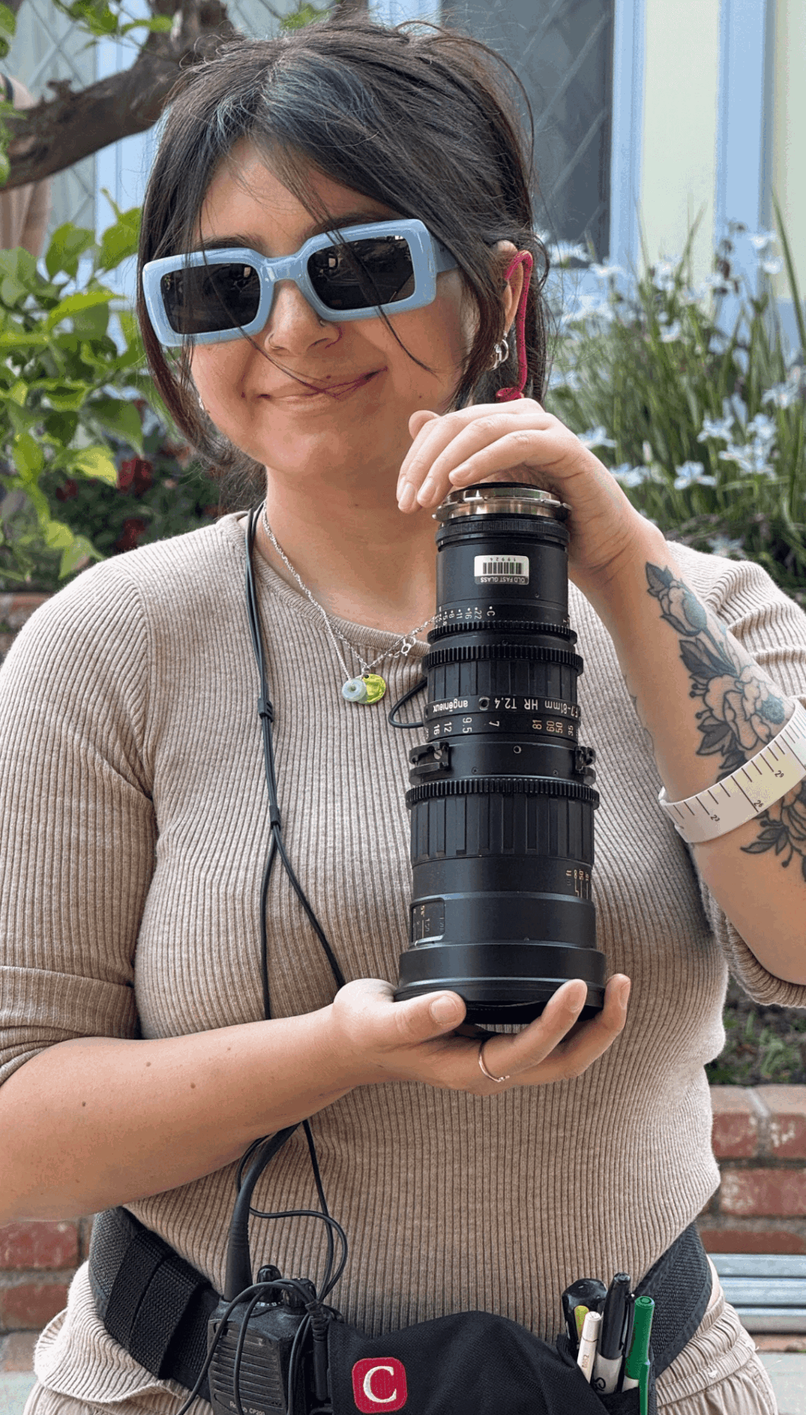 Person holding a camera outdoors, wearing sunglasses and a beige top, with a garden background.