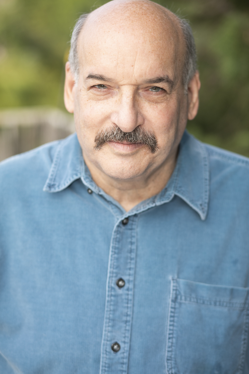 Portrait of an older man with a mustache, wearing a denim shirt outdoors, smiling, with blurred greenery in background.