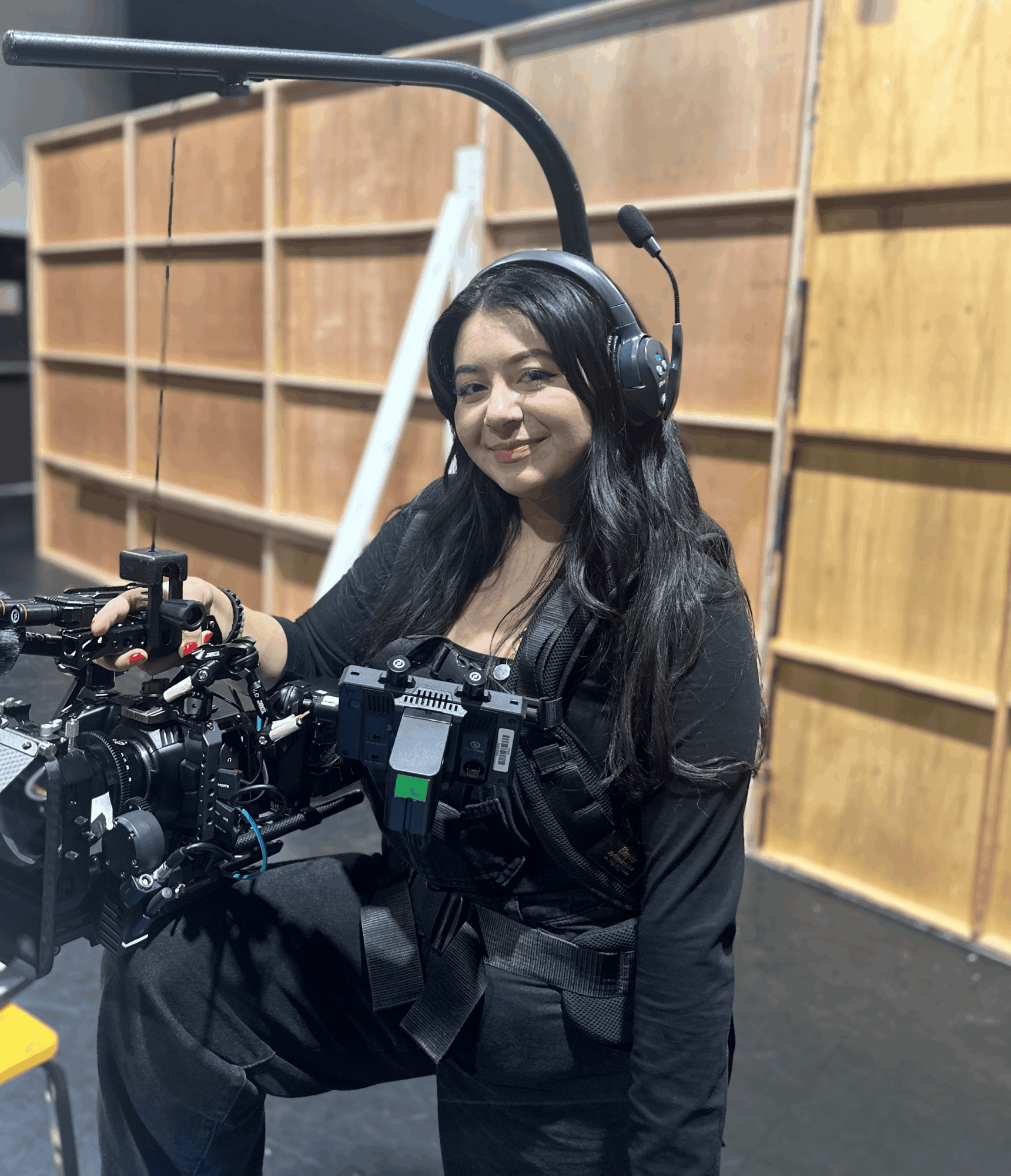 Woman with long dark hair wearing headphones, sitting at camera equipment in a room with wooden shelves.